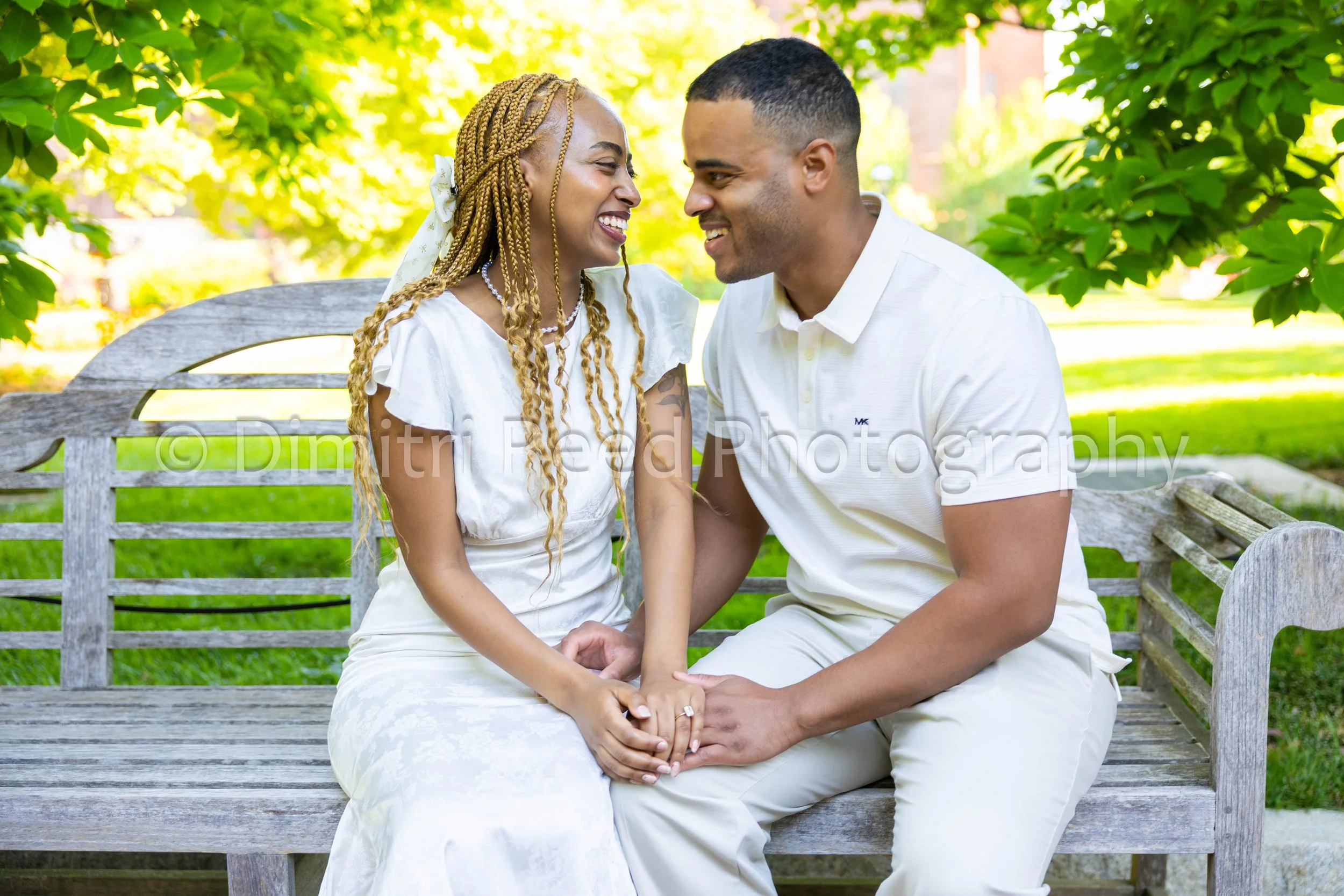 A happy couple sitting close together on a wooden park bench, holding hands and smiling at each other under a canopy of green leaves.