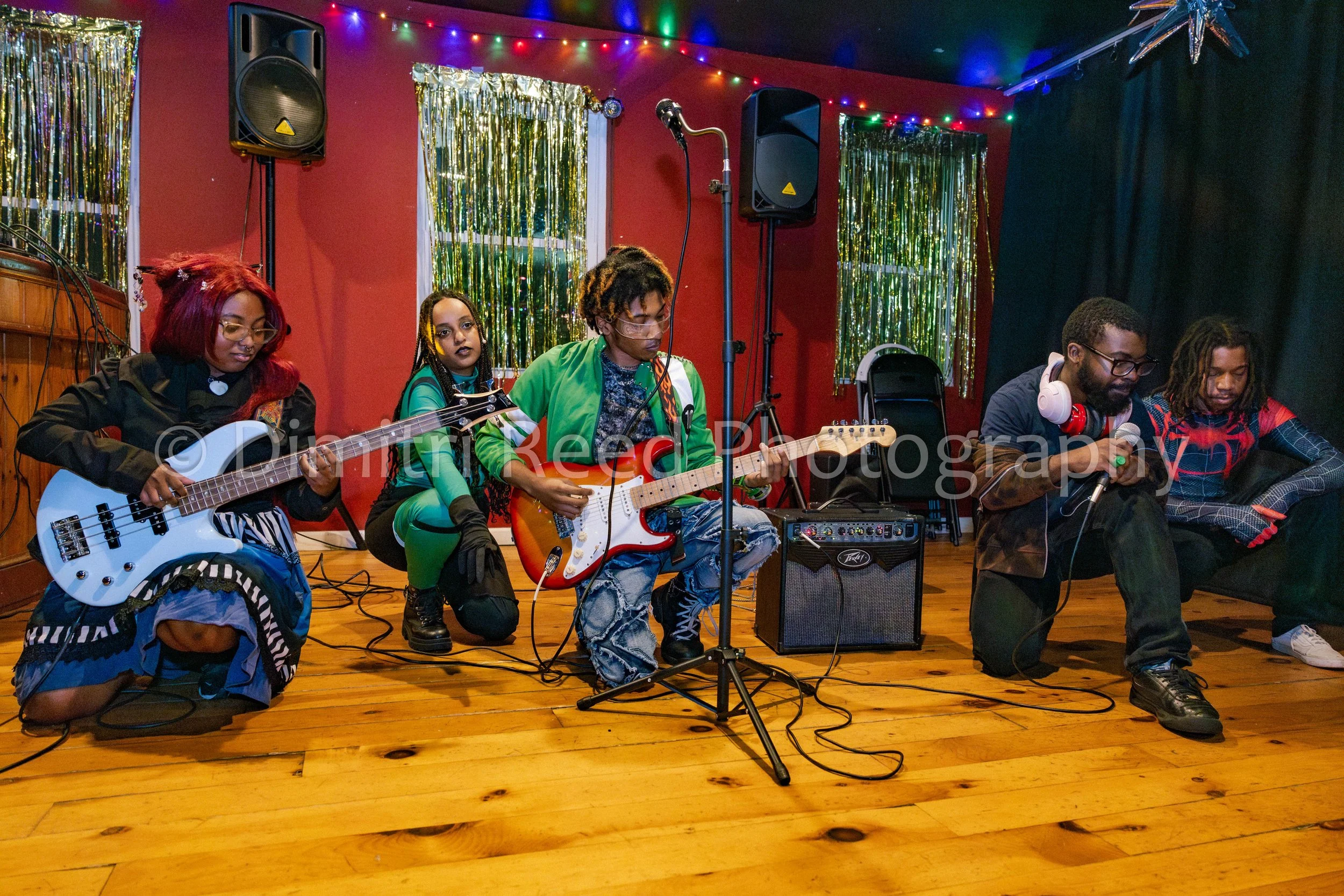 A group of five musicians playing guitars and singing on a stage decorated with multicolored string lights and gold tinsel curtains, with a black curtain on the right side.