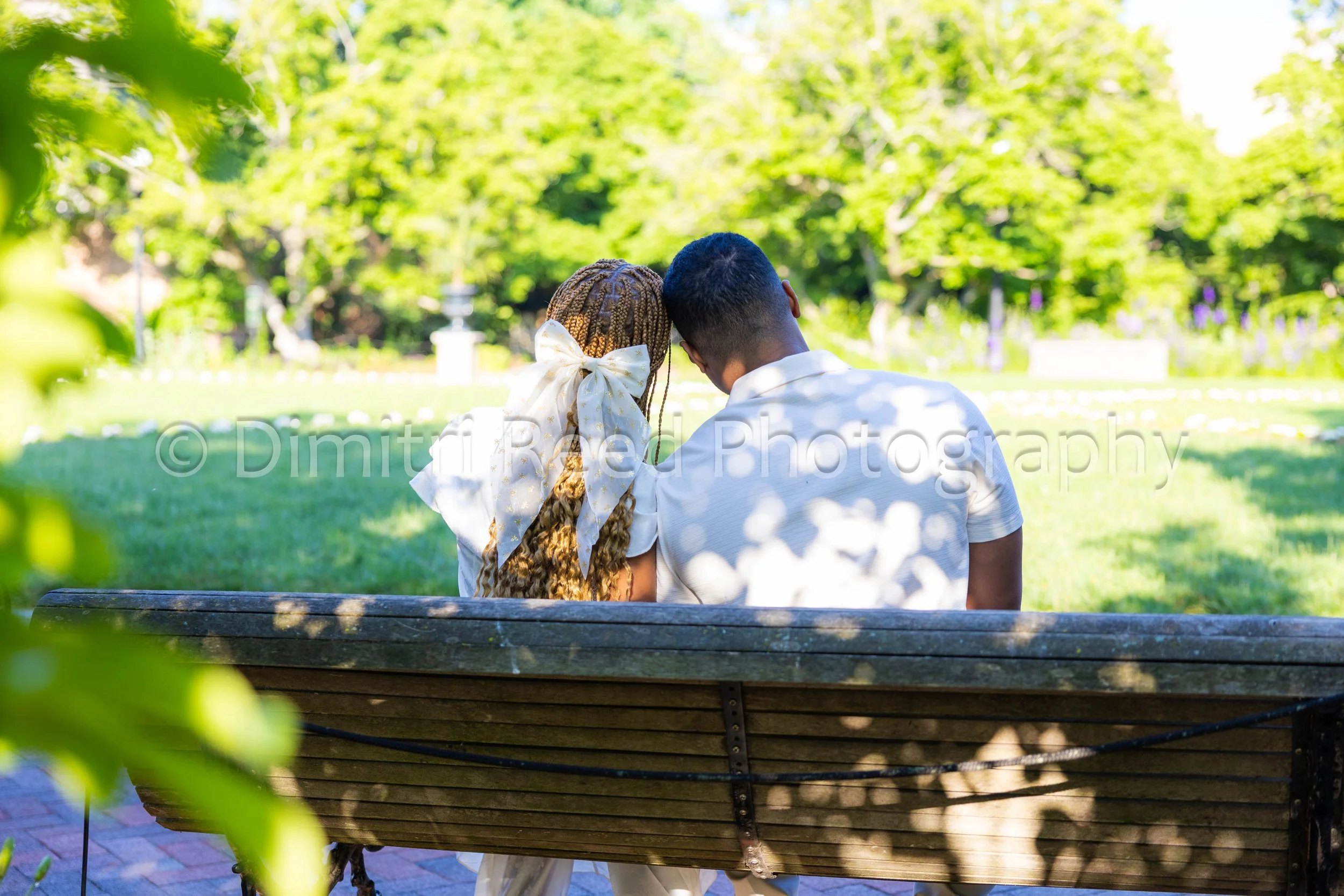 A couple sitting closely on a park bench with greenery and trees in the background.
