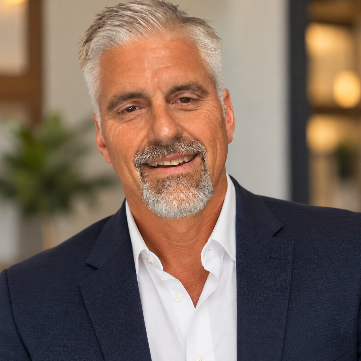 Close-up of a smiling middle-aged man with gray hair and beard, wearing a dark suit and white shirt, in an indoor setting with blurred background.