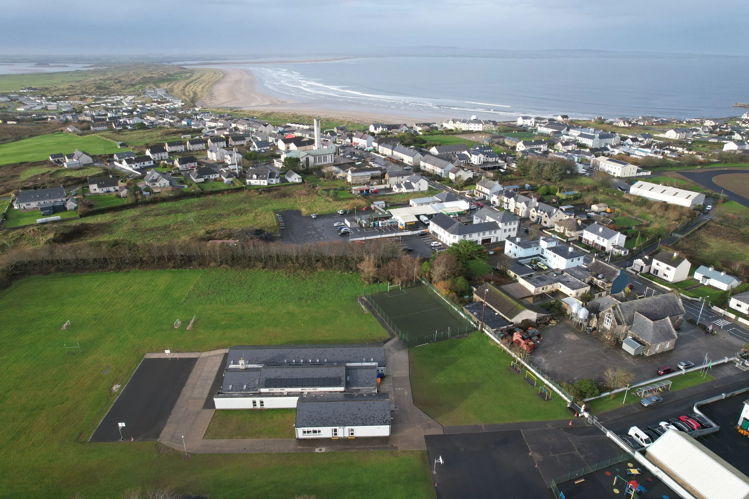 Aerial view of a coastal town with houses, sports fields, and parking lots near the ocean with waves and a sandy beach.