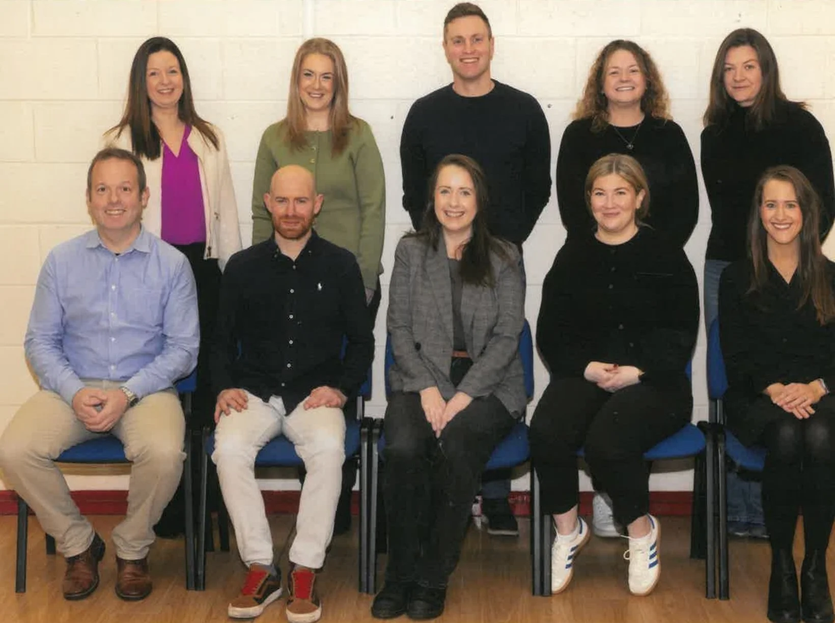 Group of ten smiling adults posed for a photo in front of a white brick wall. Some are seated on blue chairs while others stand behind them. The group consists of five women and five men, dressed in casual and business casual attire.