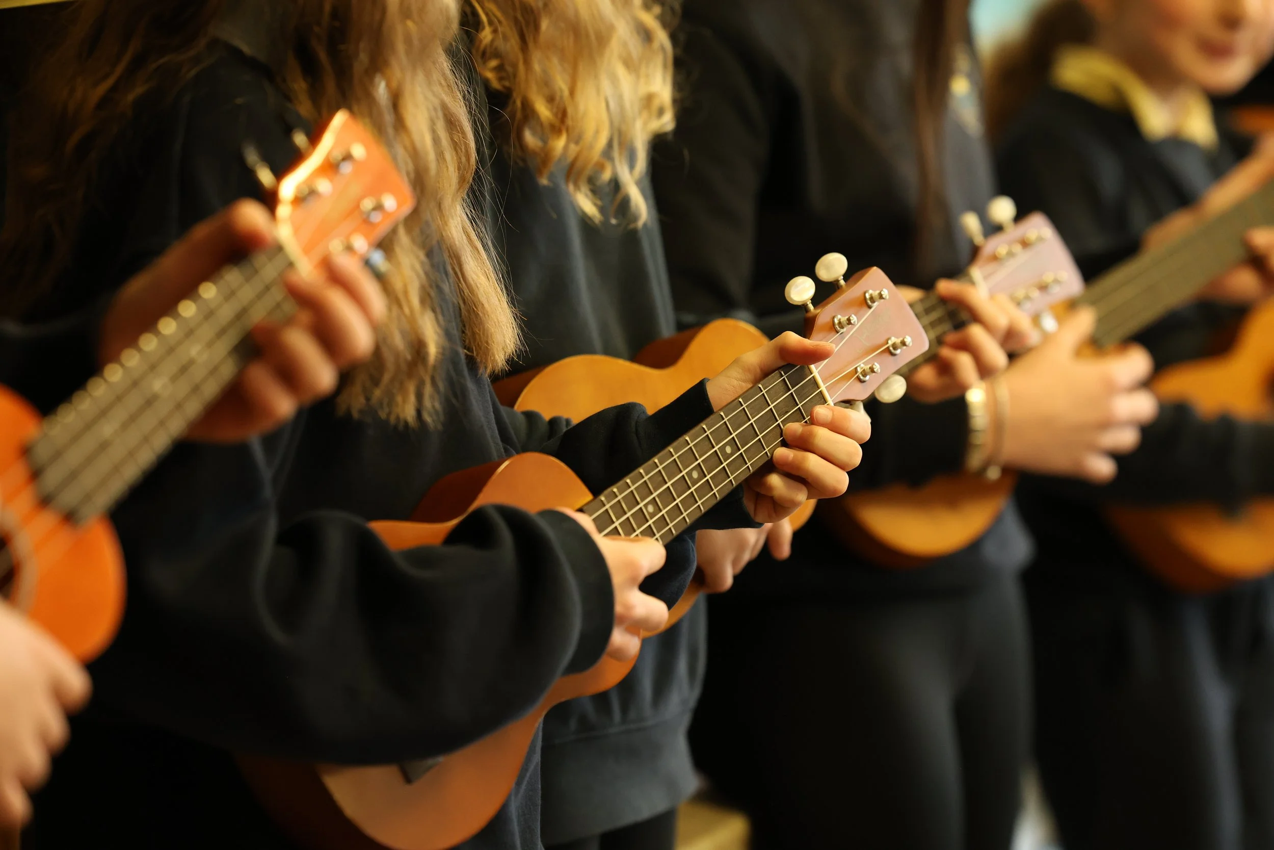 Children standing side by side, playing ukuleles in a group, wearing dark clothing, some with yellow accents, and focusing on their instruments.