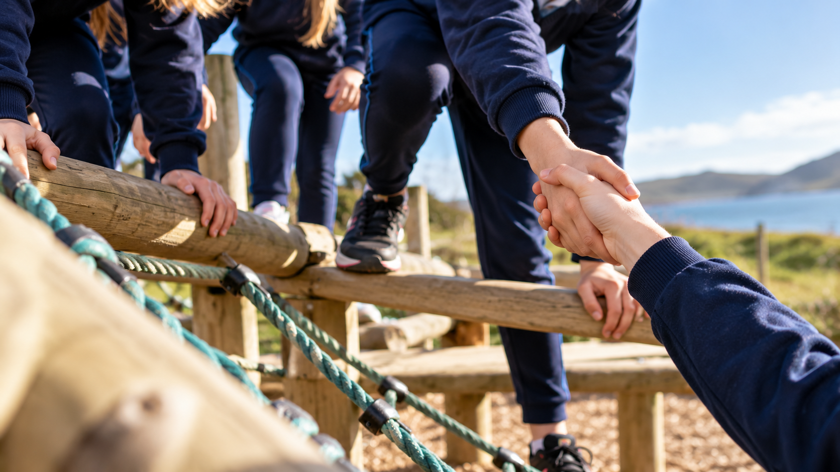 Children on a playground bridge with a person assisting, holding their hands for support on a sunny day near the coast.