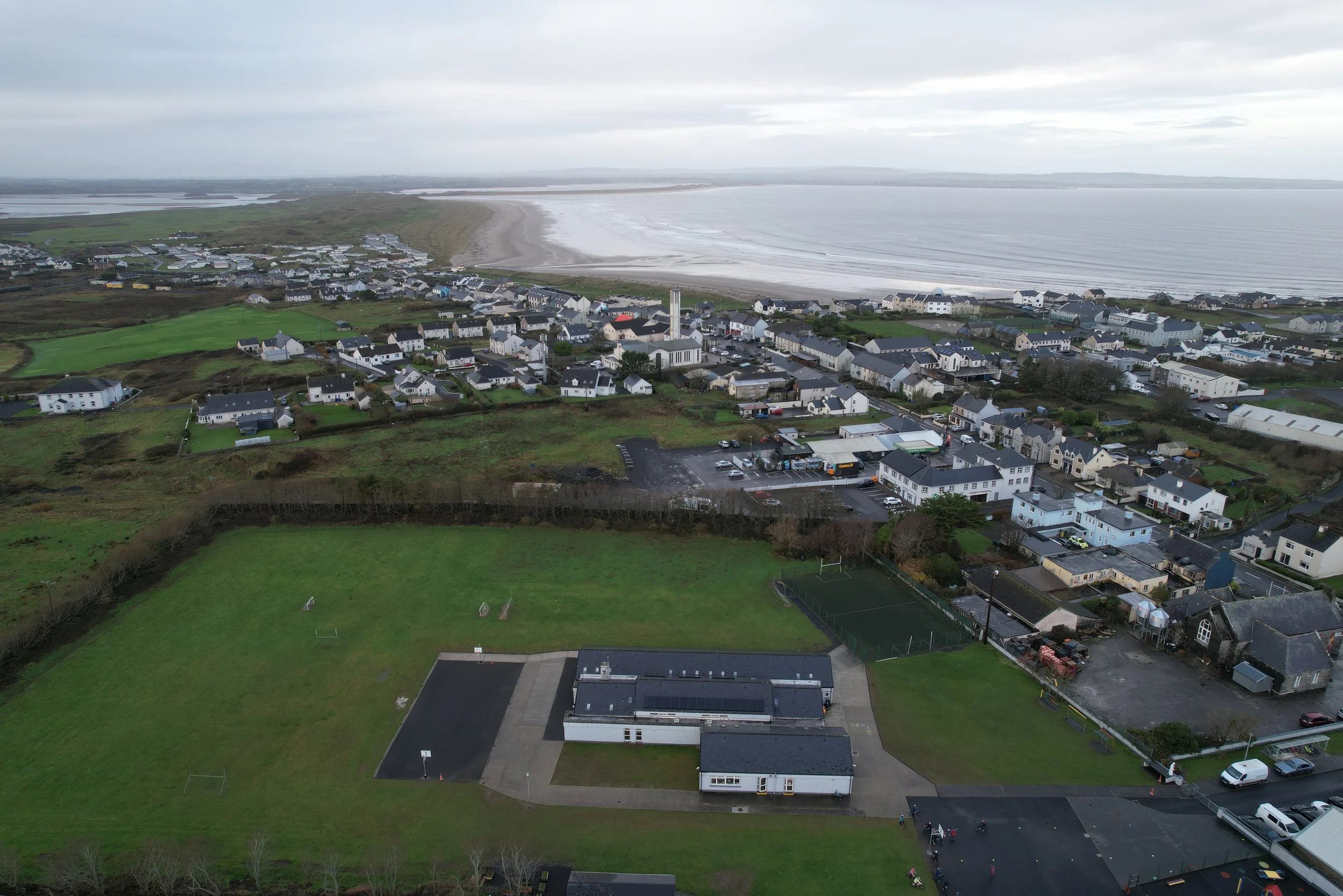 Aerial view of a coastal town with houses, a school, green sports field, and nearby beach and ocean under a cloudy sky.