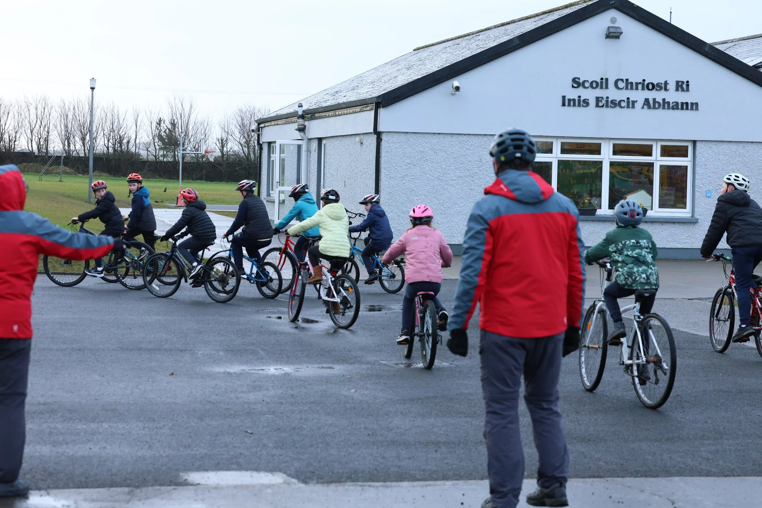 Children and adults riding bicycles outside a school building on a cloudy day, with a sign in a foreign language on the building.