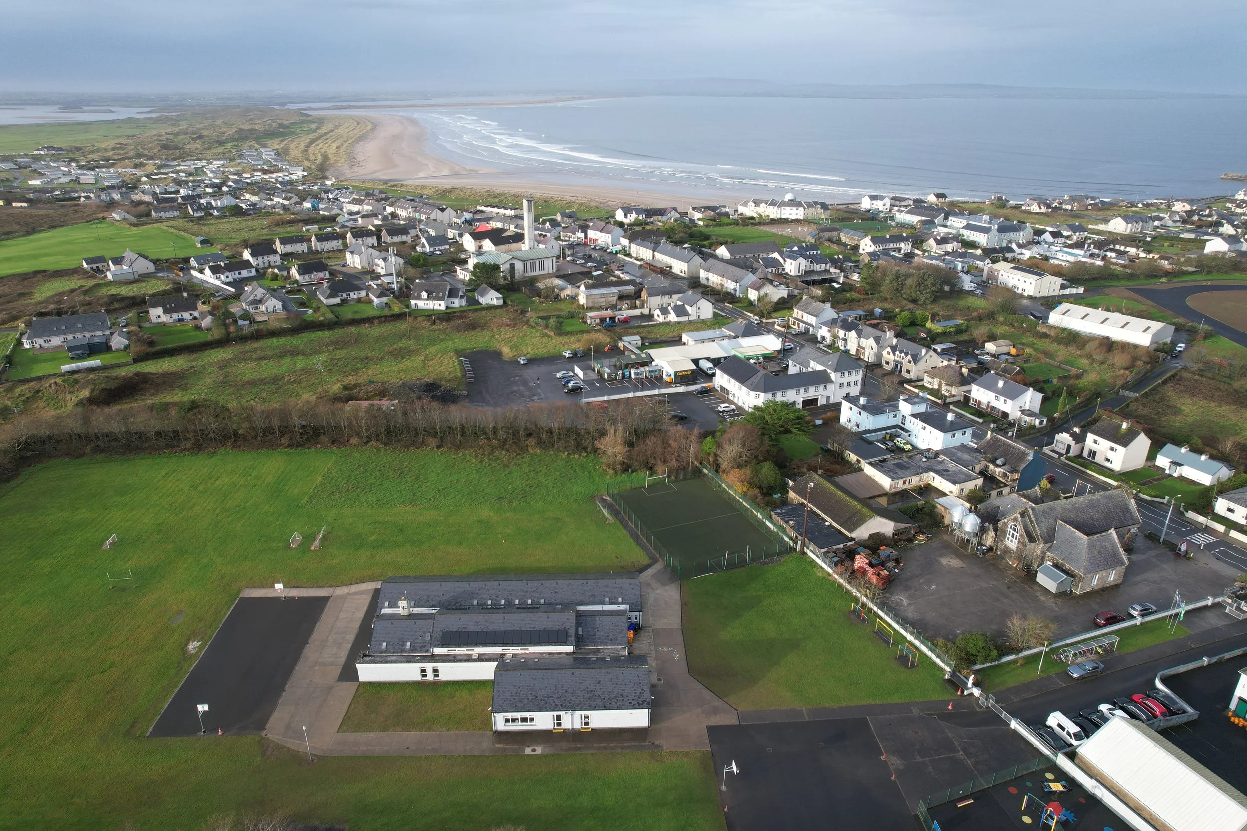 Aerial view of a coastal town with houses, green fields, and a beach along the shoreline with gentle waves, under cloudy sky.