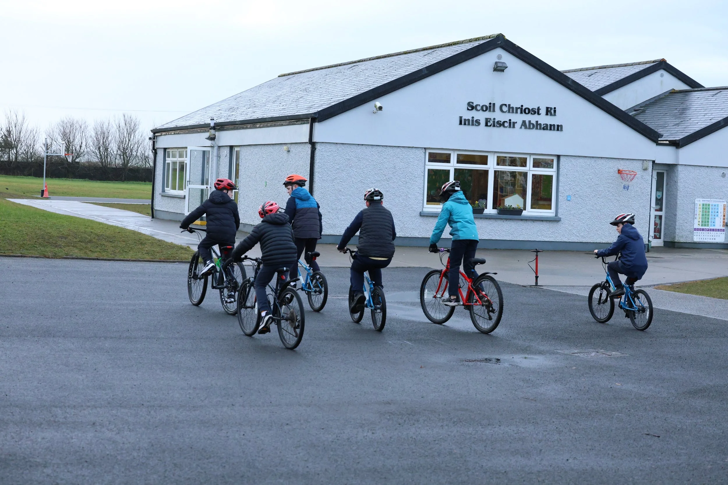 A group of children riding bicycles outside a building with a sign in Irish that reads 'Scoil Christ Ri Inis Eiscir Abhann'. The children are wearing helmets and jackets, and they are riding on a wet paved area near a grassy field, with leafless trees in the background.