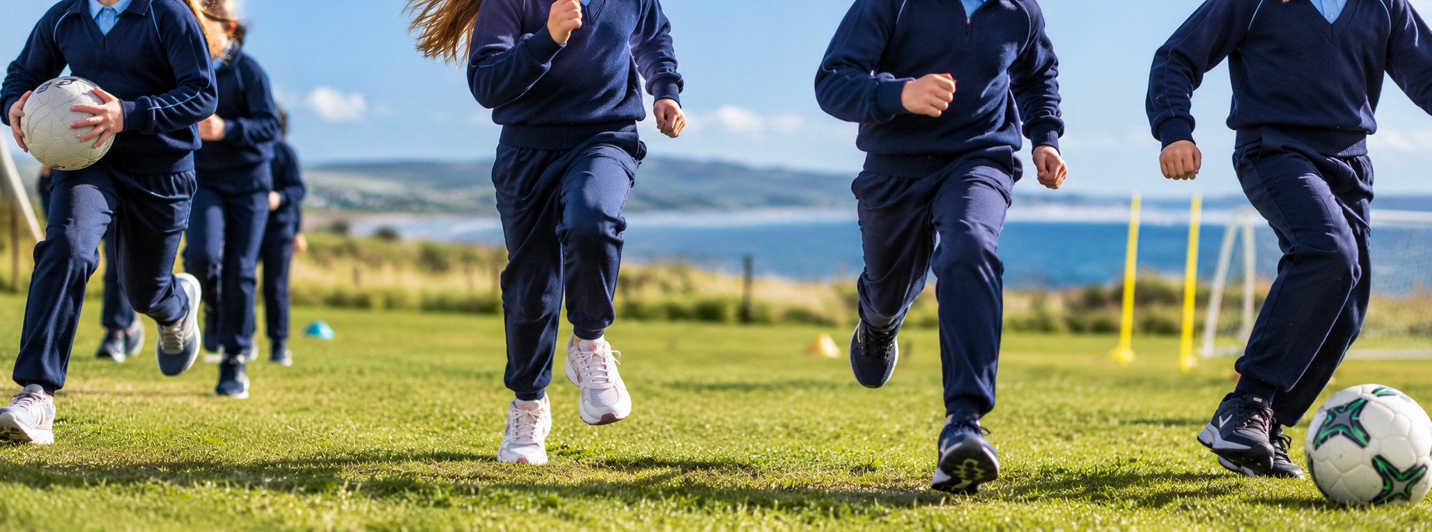 Children in navy blue sports uniforms running on a soccer field with a ball, during a sunny day with a scenic water view in the background.