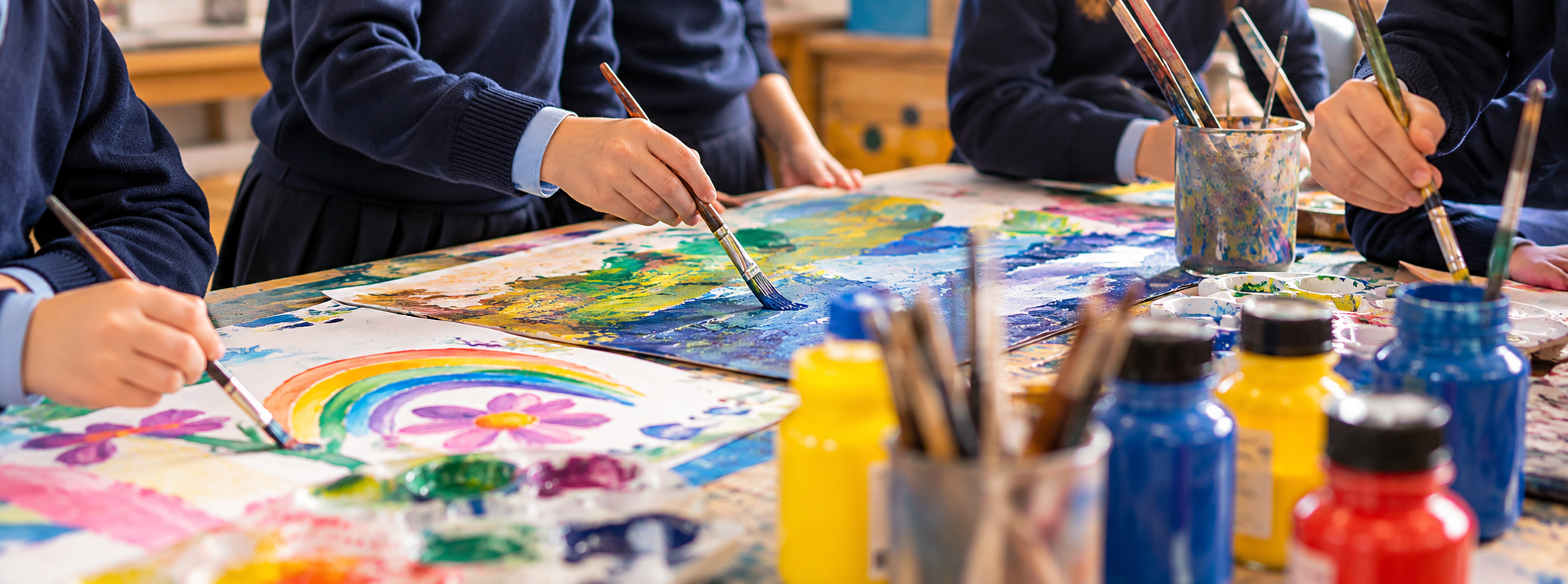 Children in navy uniforms painting colorful pictures at a table covered with paints and brushes.