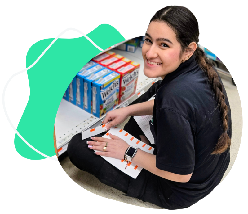 A young woman with long brown hair, wearing a black shirt and a smartwatch, is seated on the grocery store floor, looking up and smiling at the camera. She is holding a clipboard and a pen, next to shelves of cereal boxes.