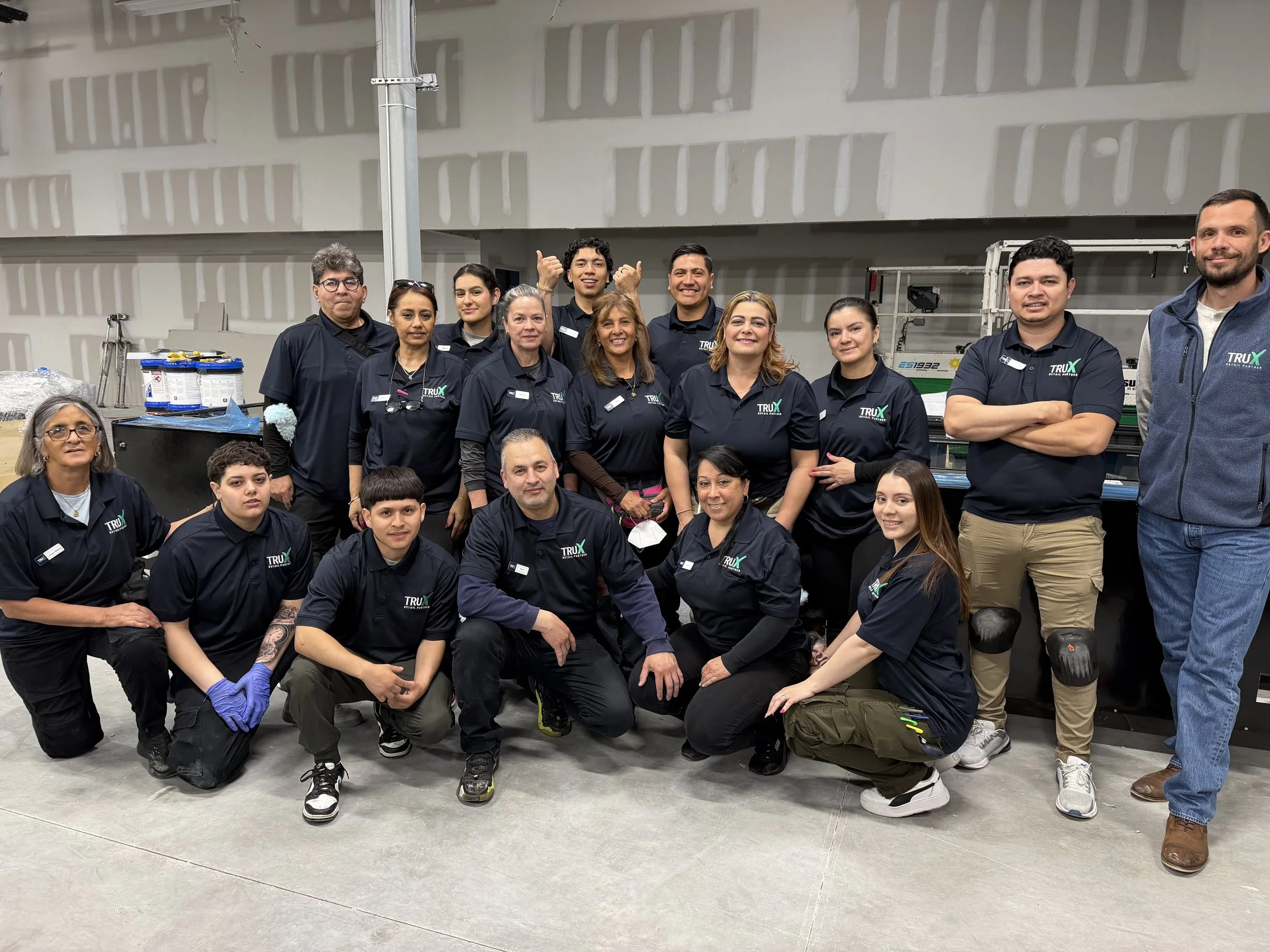 A group of diverse people posing for a photo in an indoor setting. They are wearing black or navy TRUX branded shirts, with some smiling, some giving thumbs up, and some kneeling or standing. The background shows a warehouse or industrial space with unfinished walls and equipment.