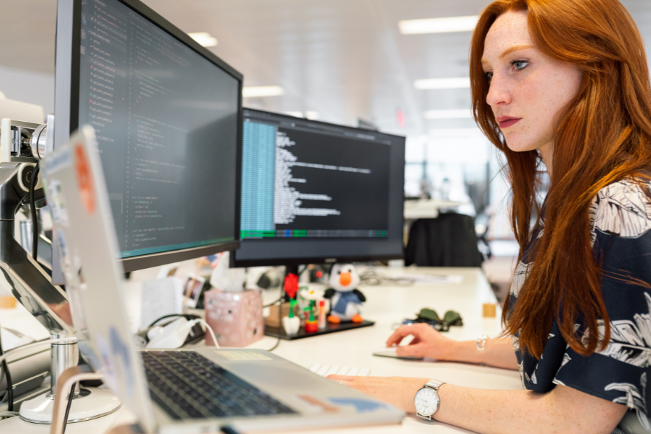 A woman with red hair working at a computer in an office setting with dual monitors displaying code and data.