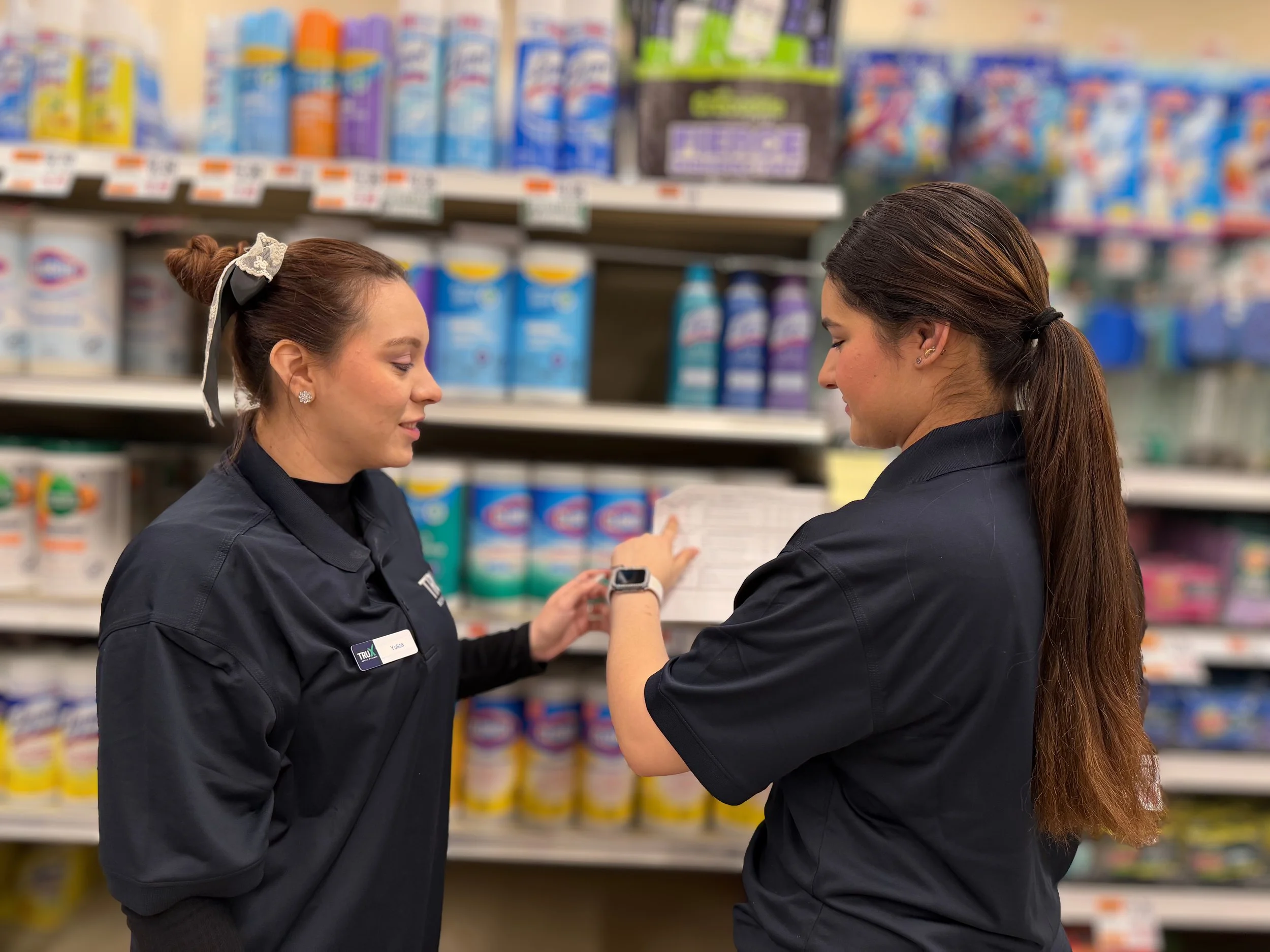 Two women with dark hair, one with a floral hair bow, are both dressed in black uniforms with name badges. They are talking and looking at a smartphone in a grocery store aisle with shelves stocked with colorful bottled drinks and other products.