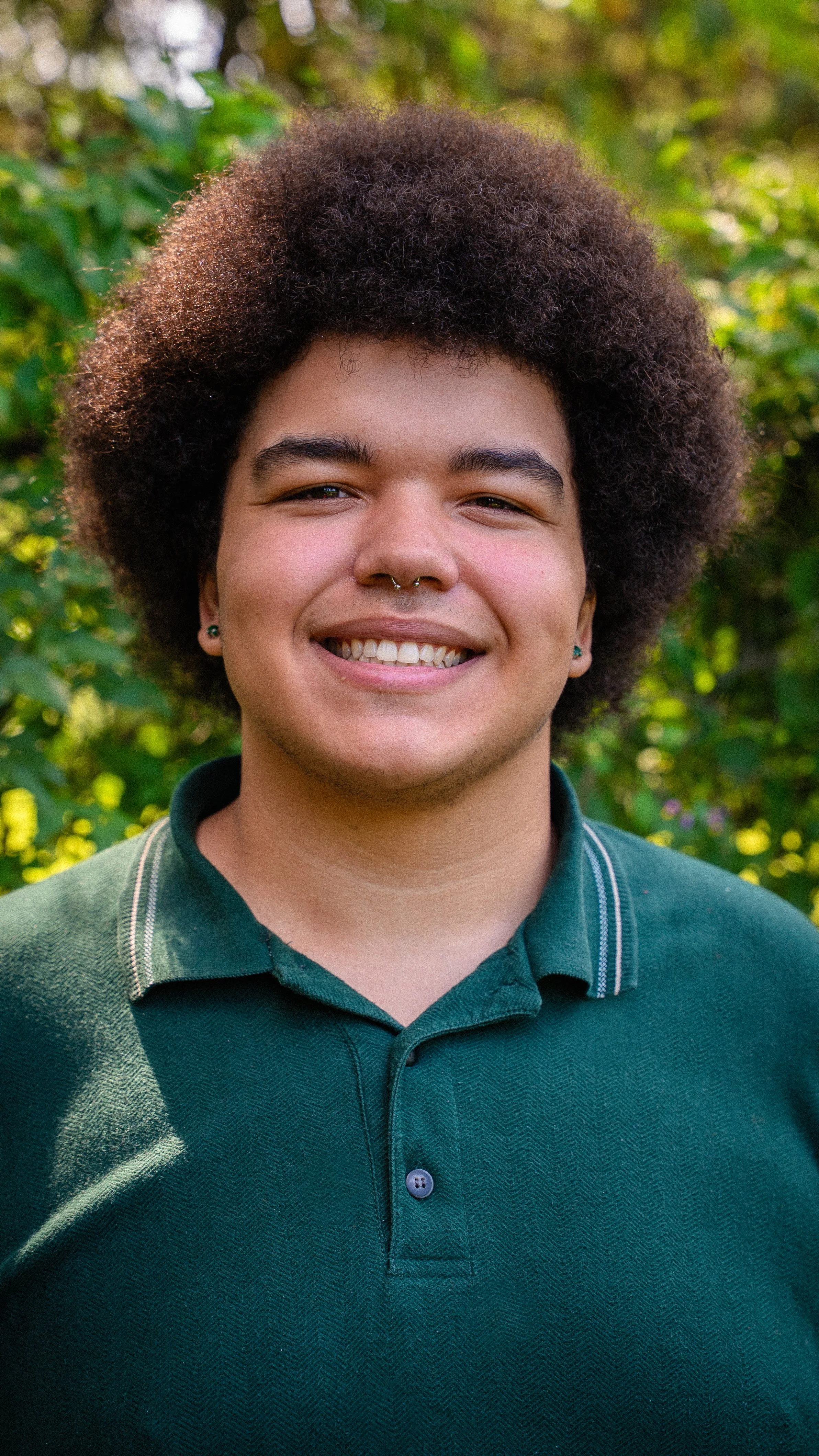 A young person with a big afro hairstyle, smiling while standing outdoors in front of greenery.