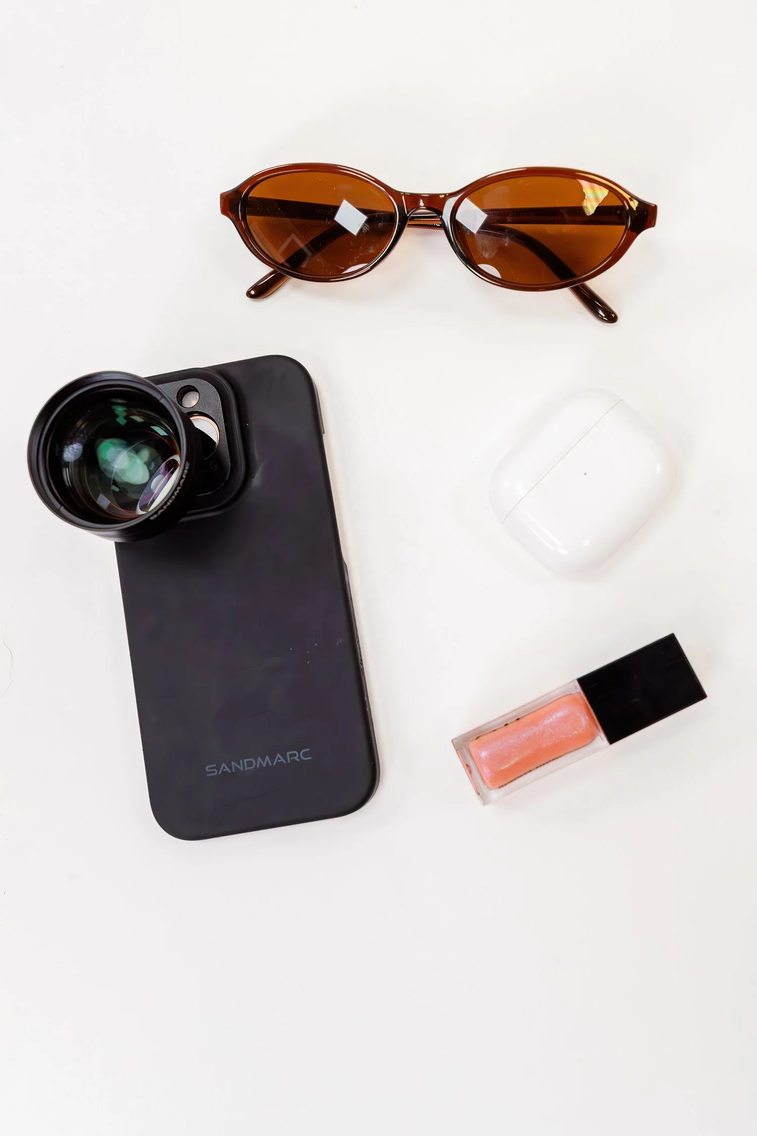 A phone and lens beside sunglasses, a white case, and lip gloss. Sandmarc product photography.