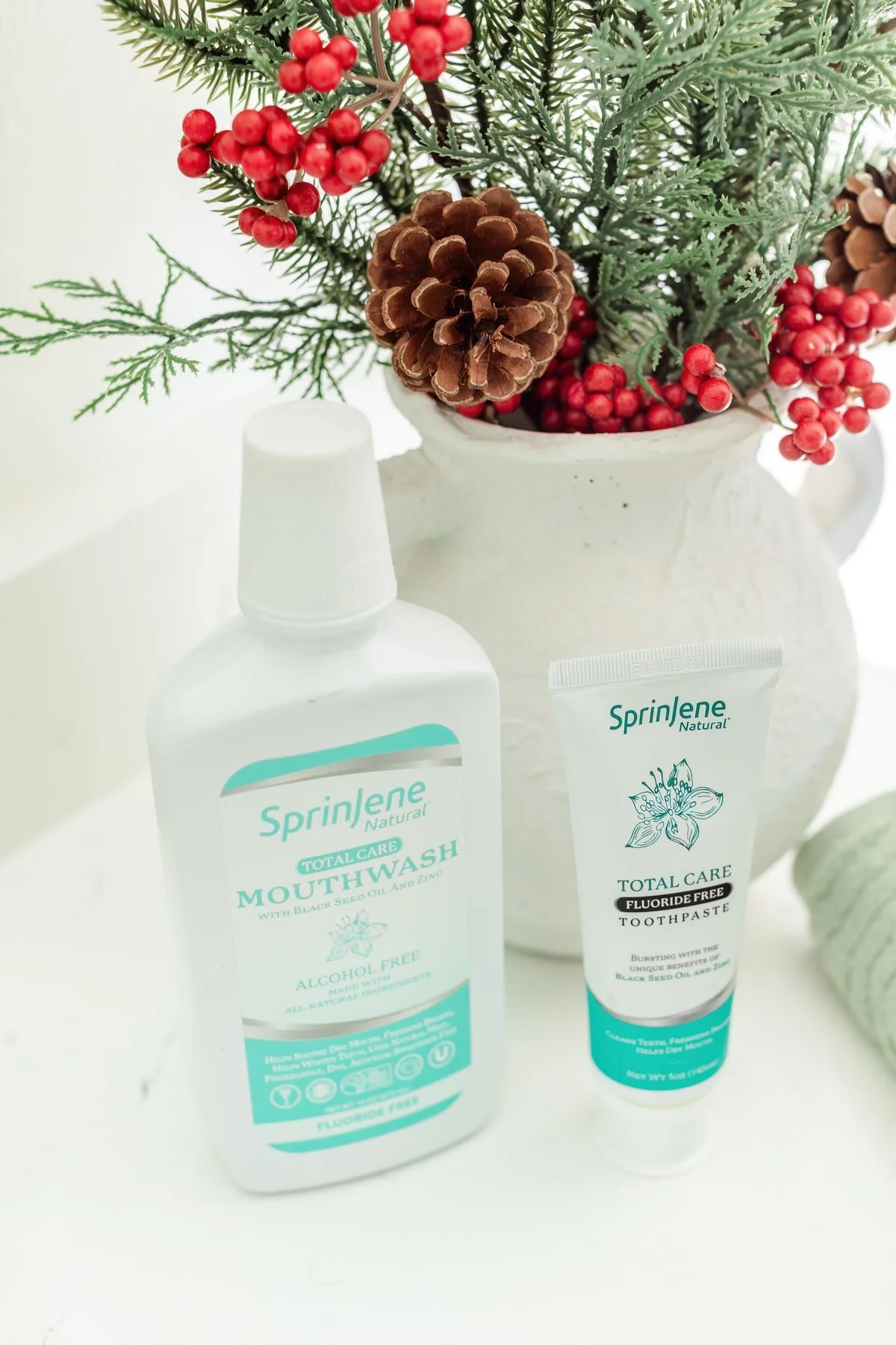 SprinJene mouthwash and toothpaste beside evergreen branches, red berries, and pine cones.