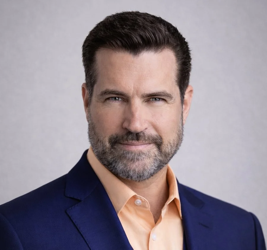 Portrait of a middle-aged man with dark hair, a beard, blue eyes, wearing a navy blazer and a light-colored collared shirt, smiling slightly against a plain background.