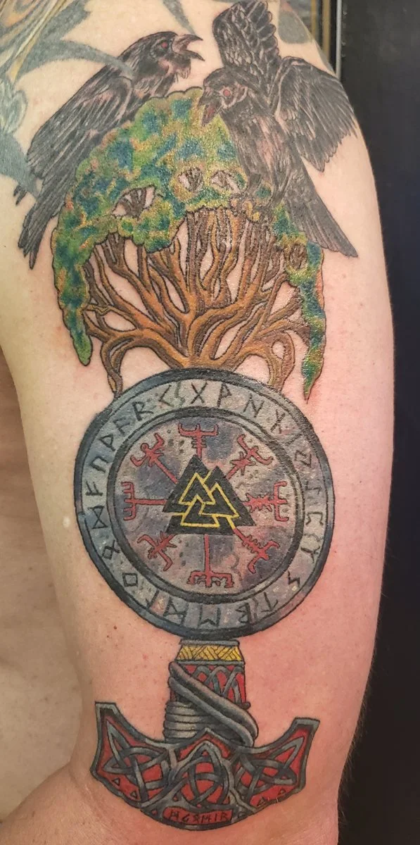 Tattoo of two black ravens perching on a tree with green foliage, above a circular symbol with runic characters and red designs, connected to a detailed red and gray axe.