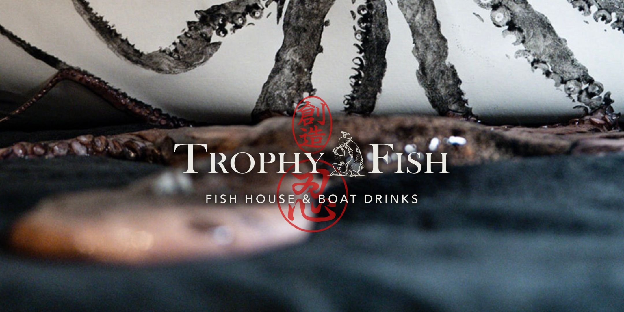 Close-up of a logo for Trophy Fish, a fish house and boat drinks establishment, placed over a background of a fish with detailed scales and fins, and a low angle view of a wooden surface.