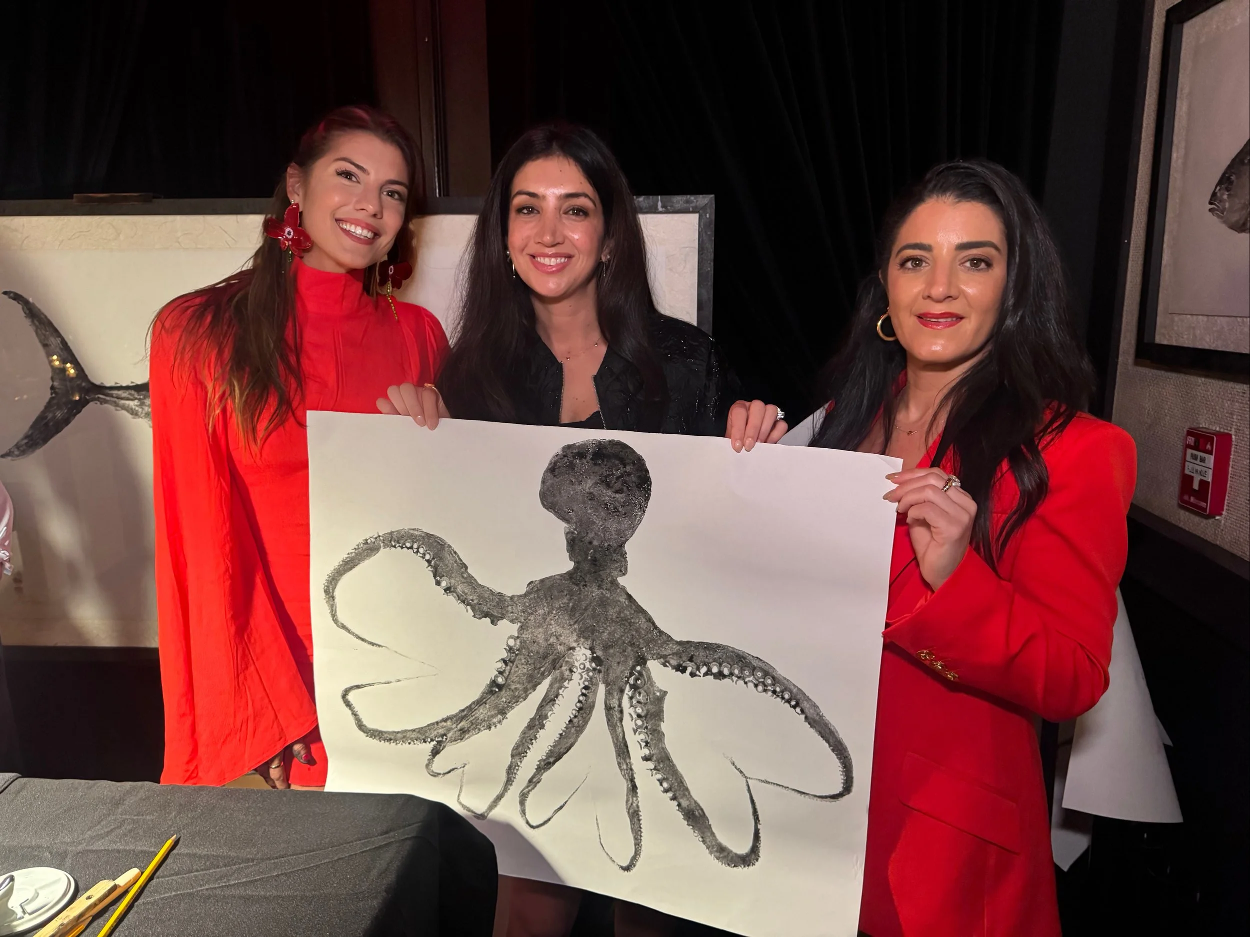 Three women holding up a painting of an octopus at an indoor event, with dark background and some tables and chairs visible.