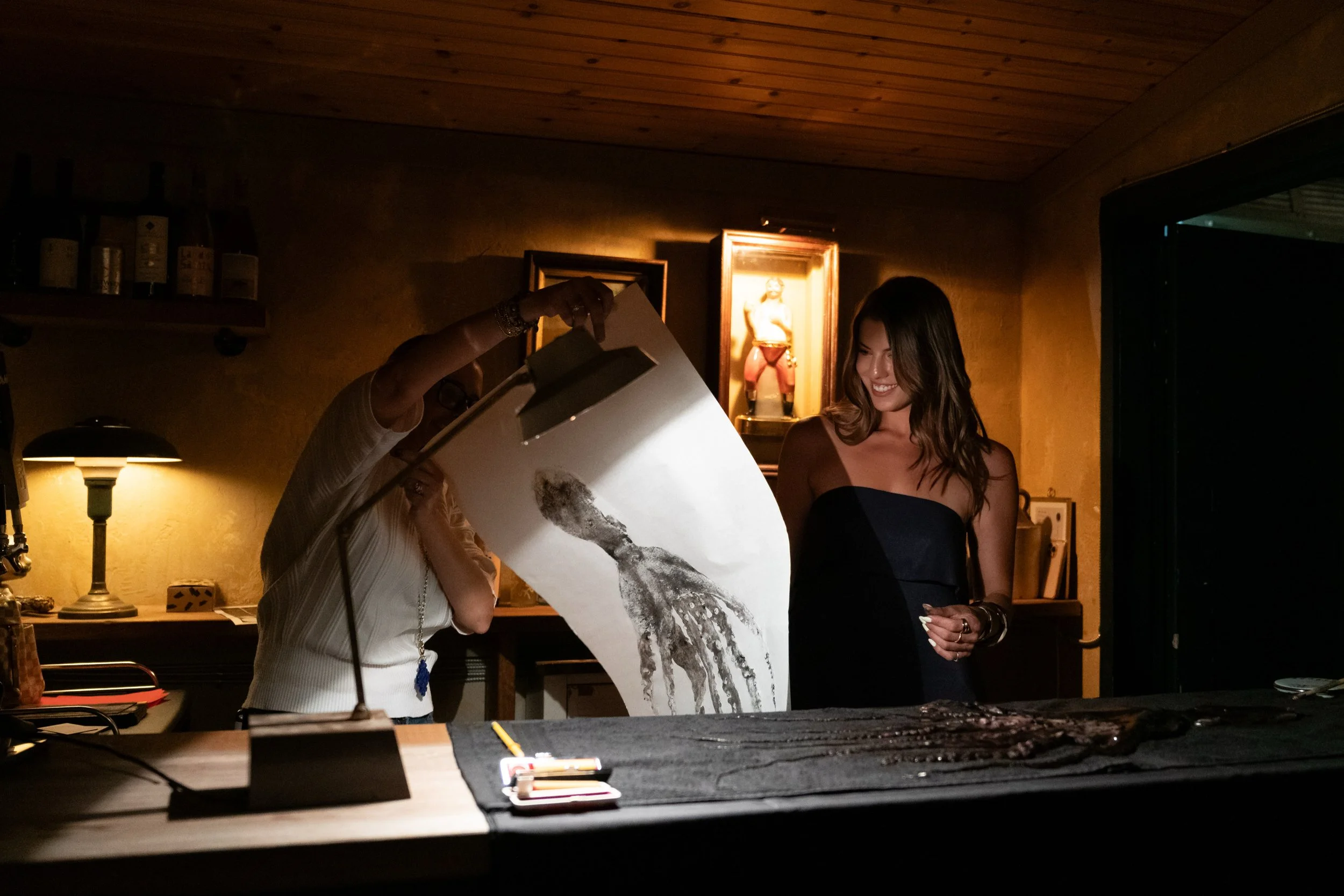 A woman in a strapless black dress laughing and smiling, standing in a dimly lit room with a person showing her artwork. The background features framed paintings and bottles on a shelf.