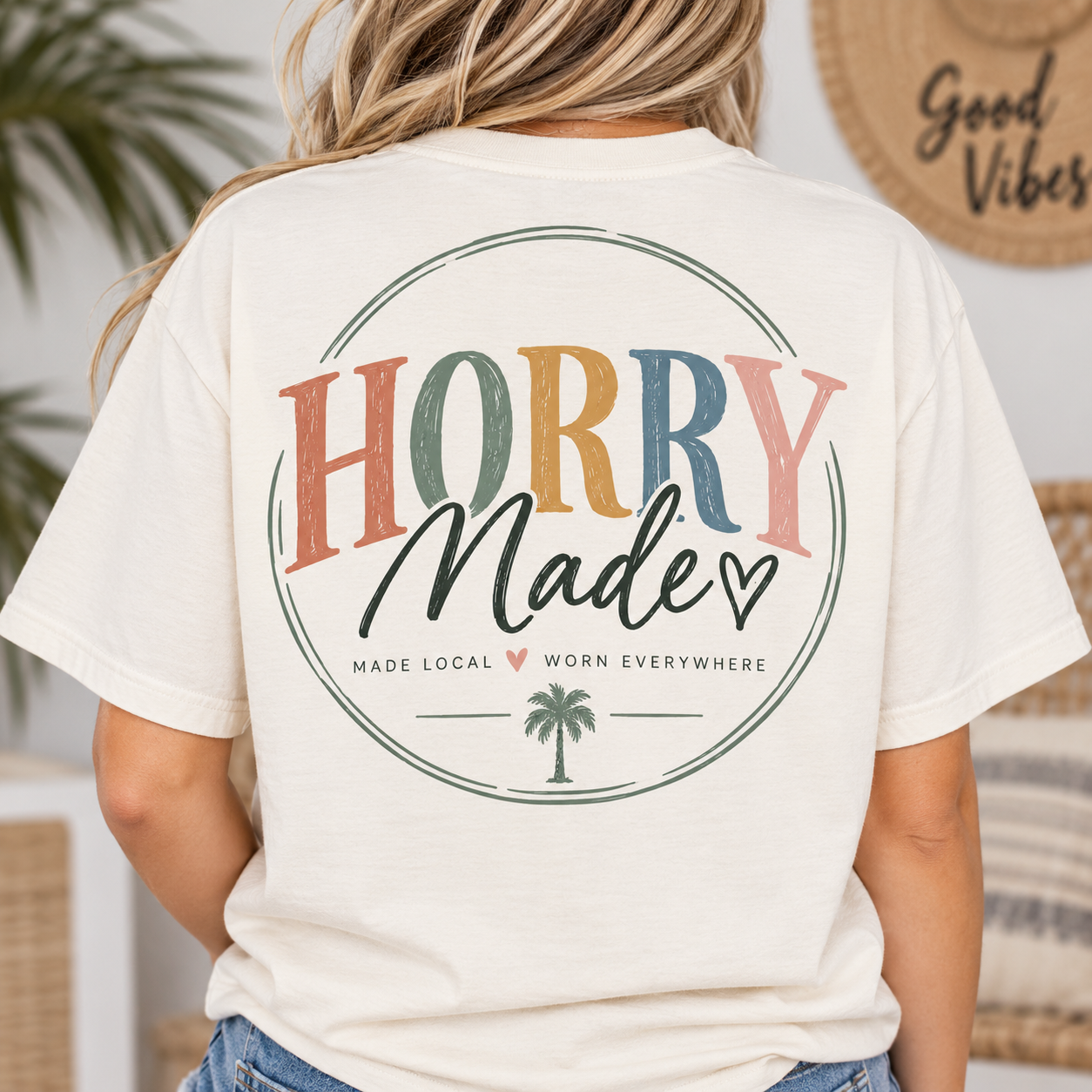 Back of a woman wearing a beige T-shirt with the text 'Hooray Made' in colorful letters, and additional text 'Made Local Worn Everywhere' with a small palm tree graphic.