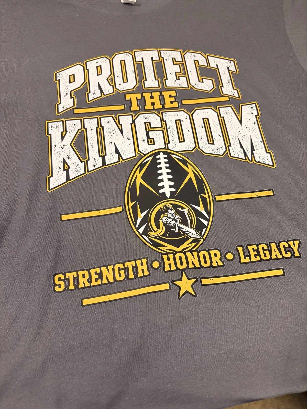 Gray shirt printed with Protect the Kingdom, a football with a knight, and Strength Honor Legacy