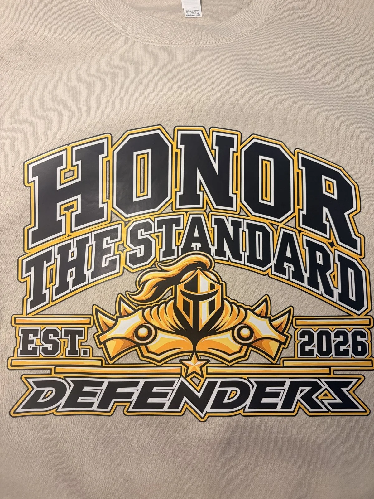 Sand tee printed with an Honor the Standard Est. 2026 Defenders knight design in gold, black and white