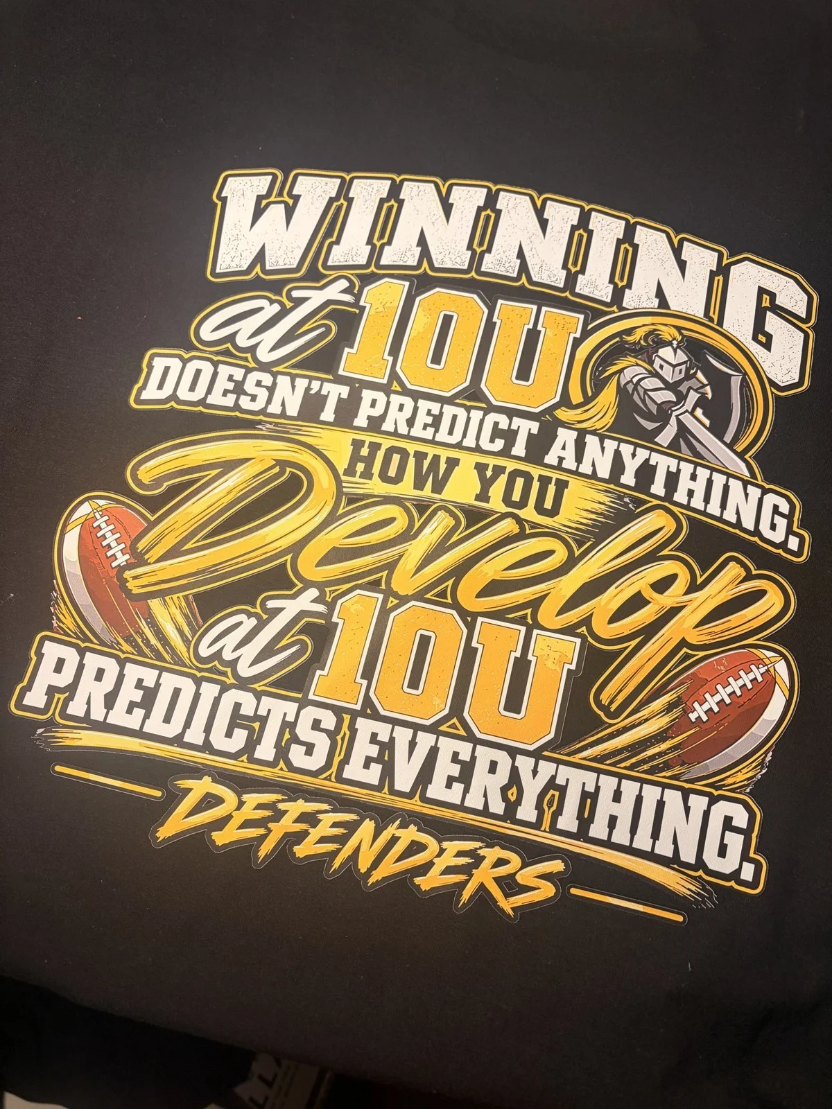 Black shirt printed with a Winning at 10U quote design with footballs and a Defenders knight