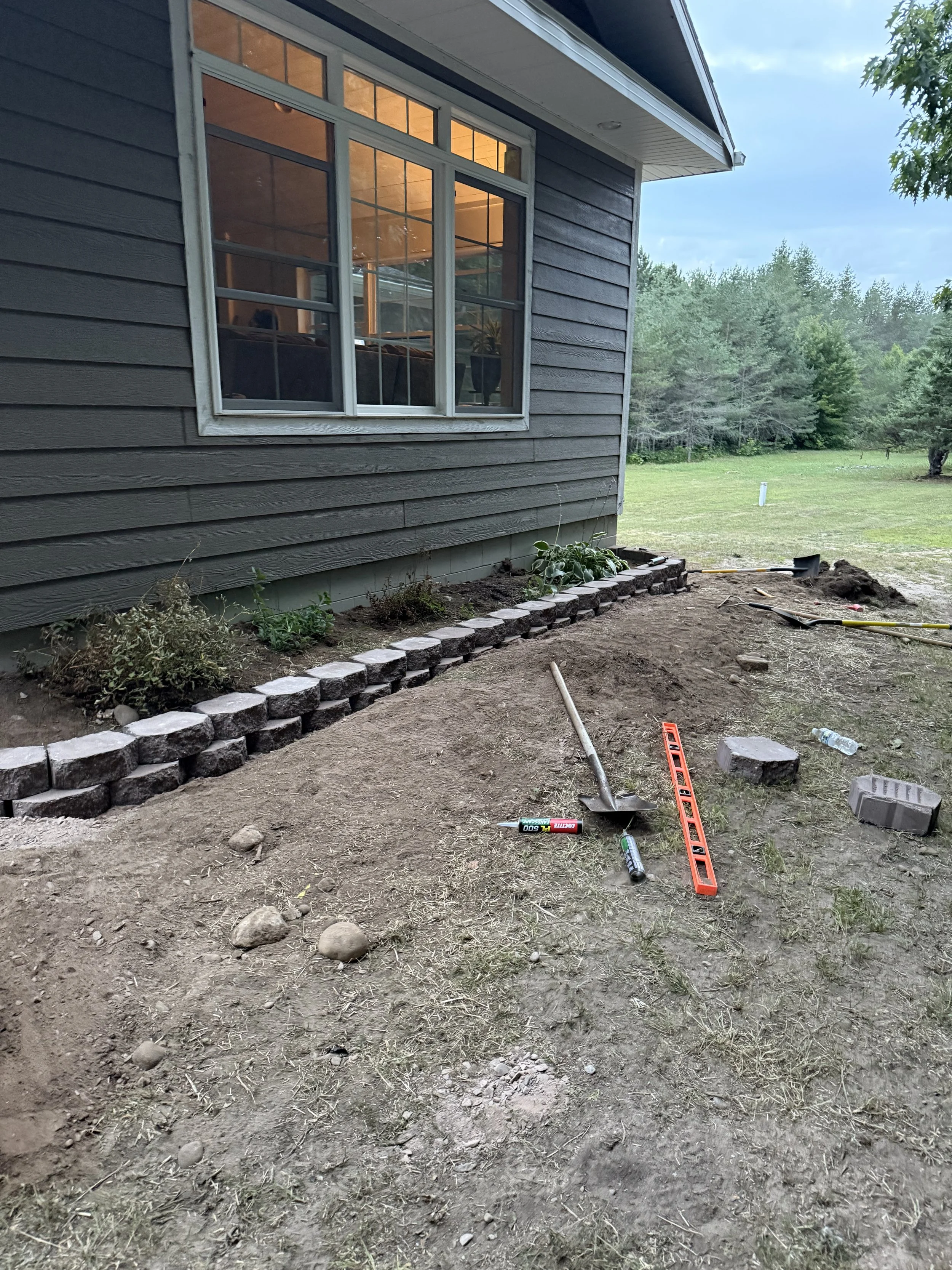 House exterior with a large window, stone garden border, exposed soil, landscaping tools, and a grassy wooded yard.