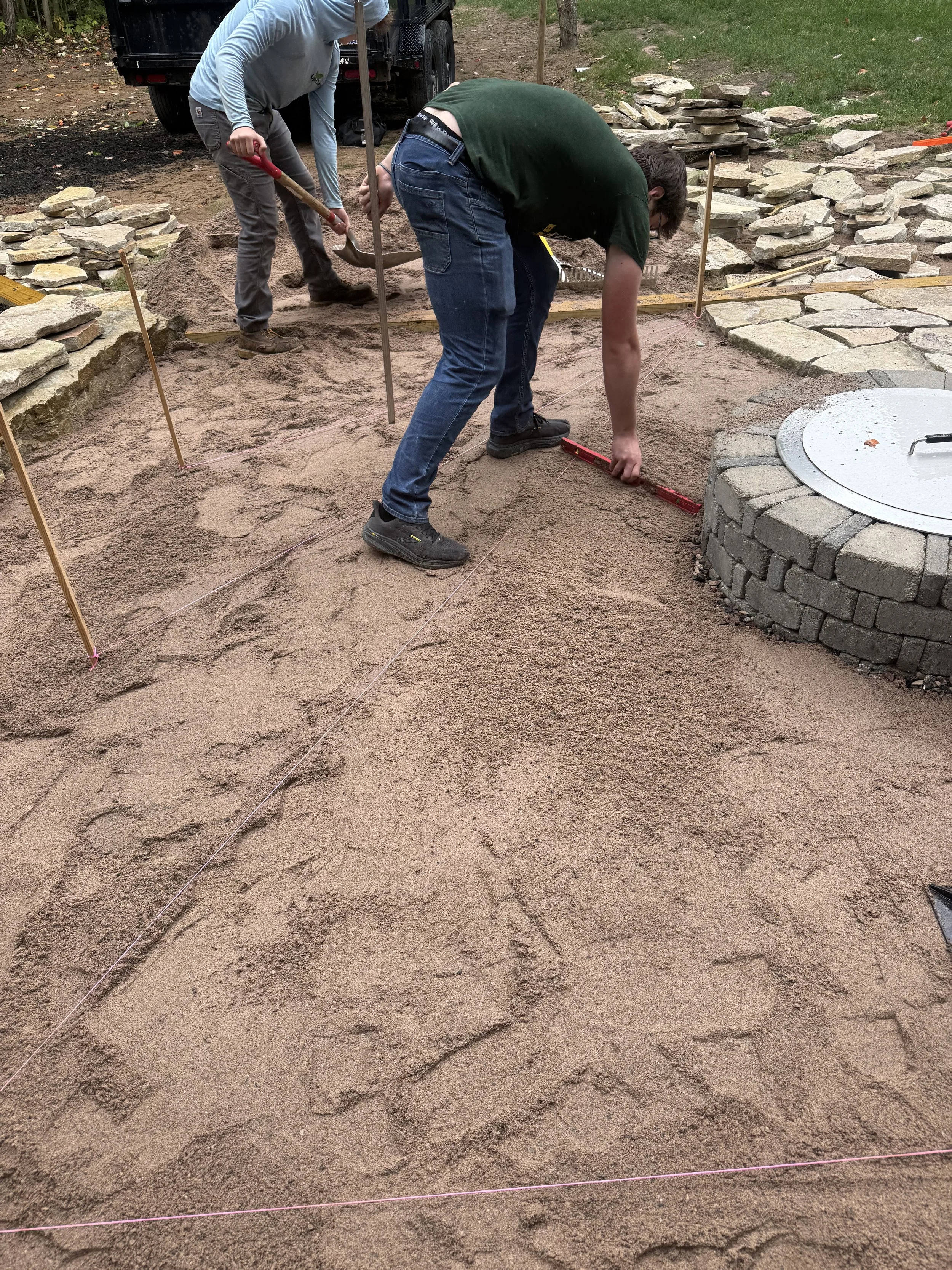 Two people working on laying out a brick pathway with string guides, one bending over and the other standing with a shovel nearby.