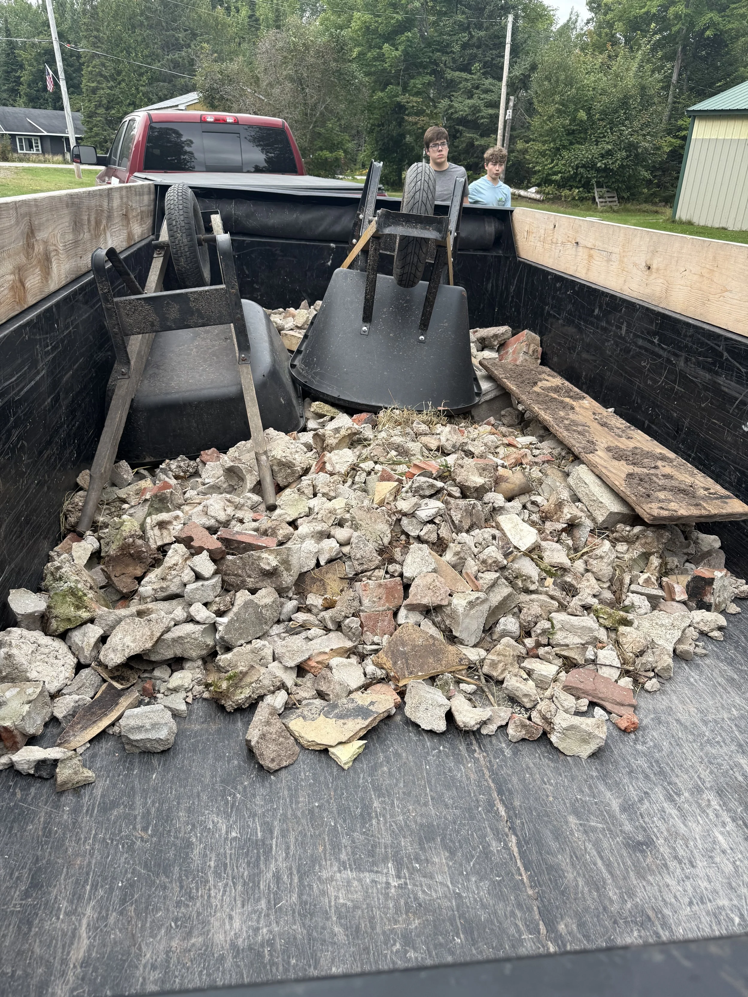 Two boys stand behind a trailer loaded with rubble, tires, and black metal equipment near a red pickup truck in a wooded residential area.
