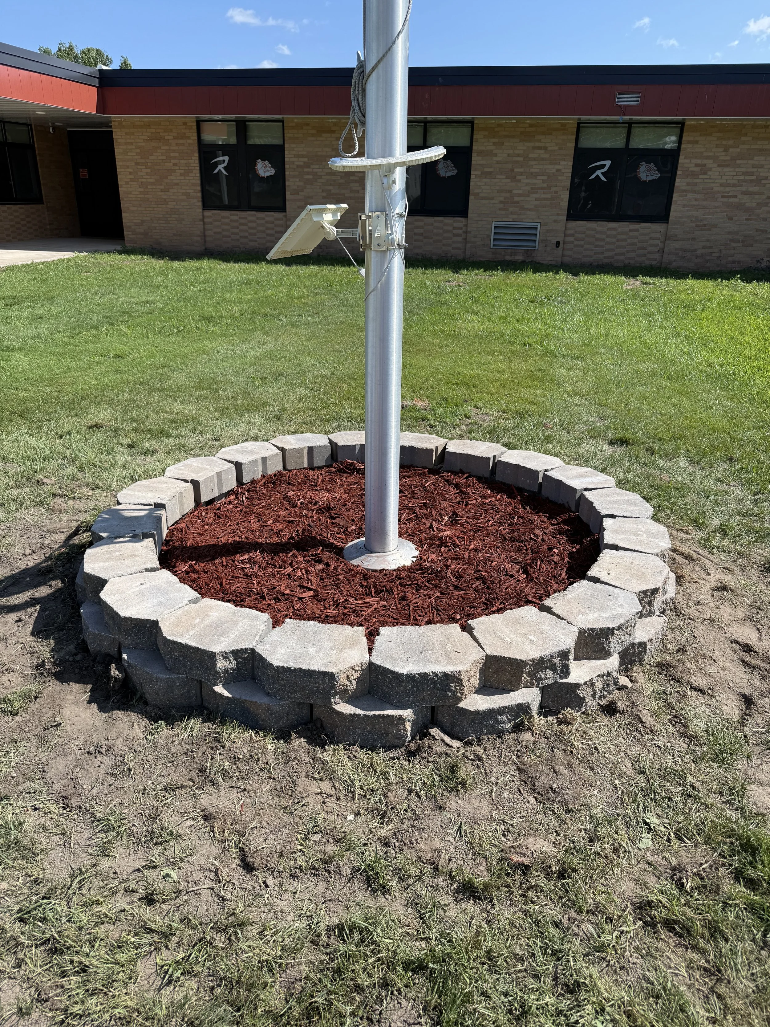 Flagpole mounted in a landscaped mulch bed outside a brick school building.