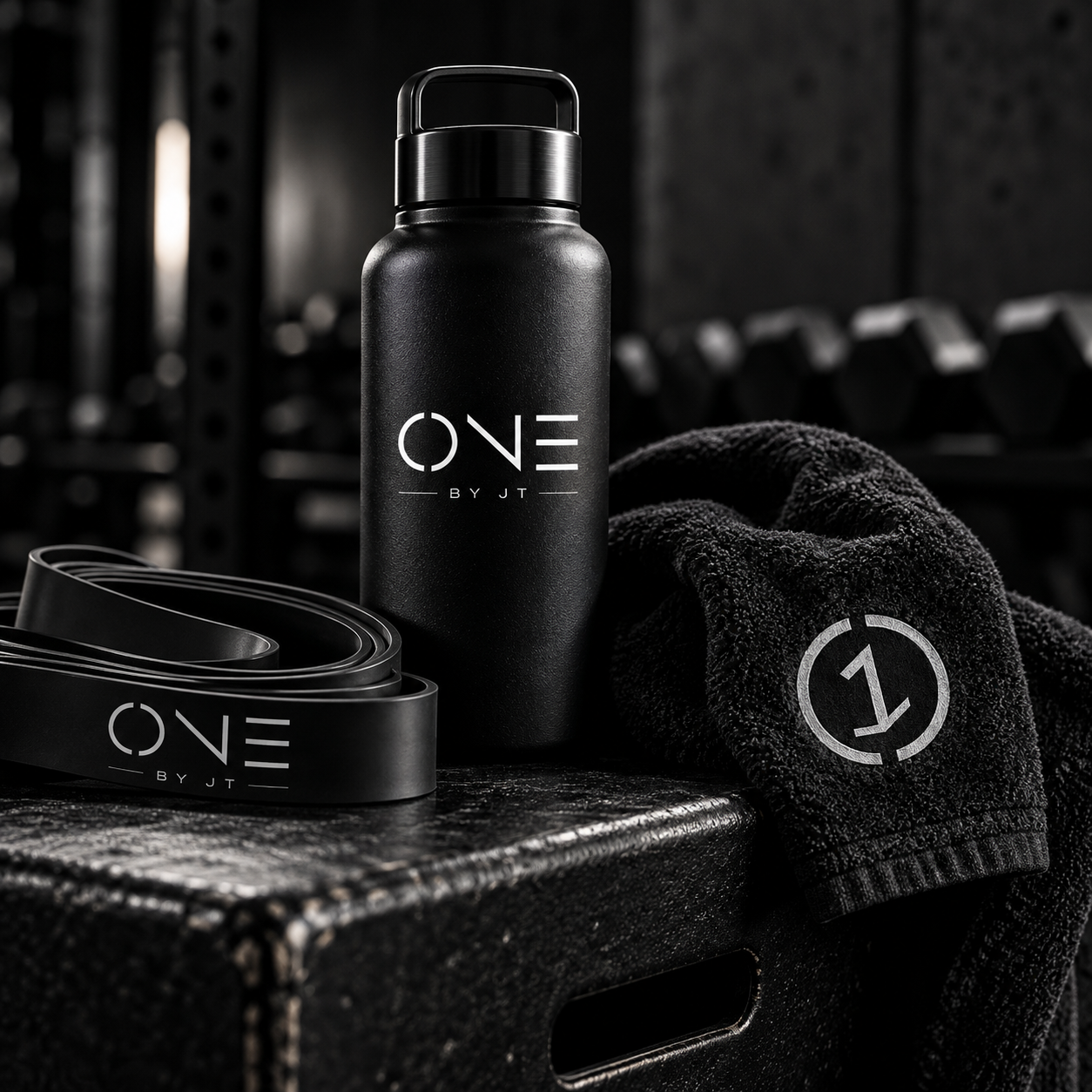A black water bottle, a black resistance band, and a black towel with a white logo, placed on a black surface in a gym setting.