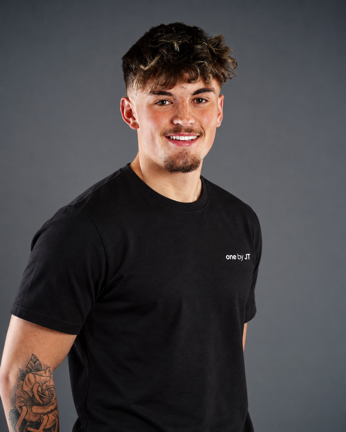 Young man with short wavy hair, light skin, and a goatee, smiling, wearing a black T-shirt with the text "one by JT" on the chest, standing against a gray background.