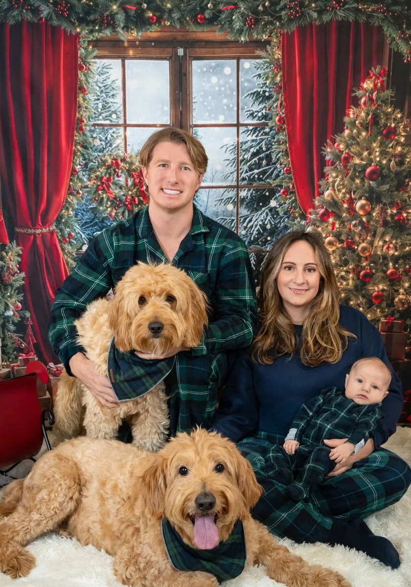 Family in plaid pajamas with a baby and two goldendoodles in a red Christmas studio set, holiday family and dog portrait