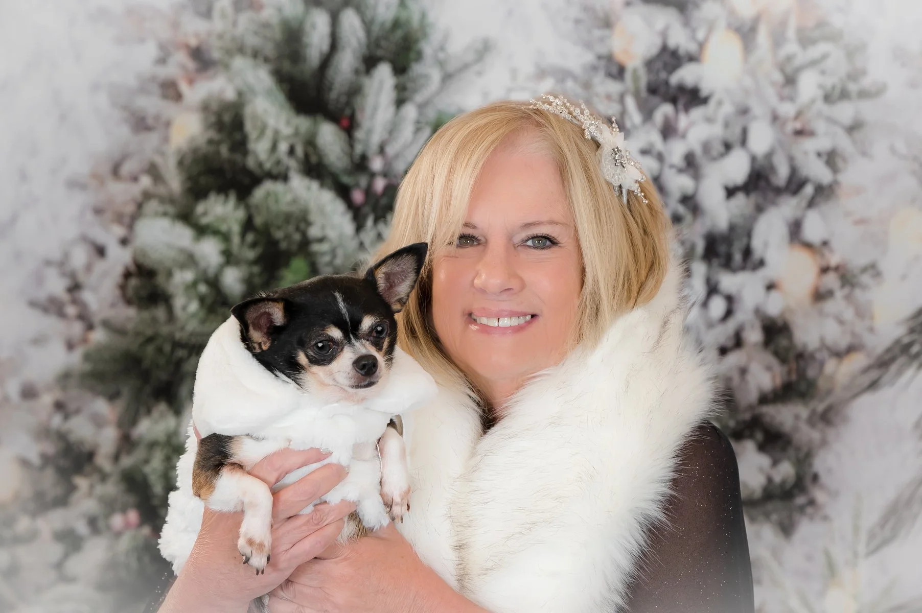 Woman in a white fur wrap holding her small dog in a snowy evergreen set, holiday portrait with pet