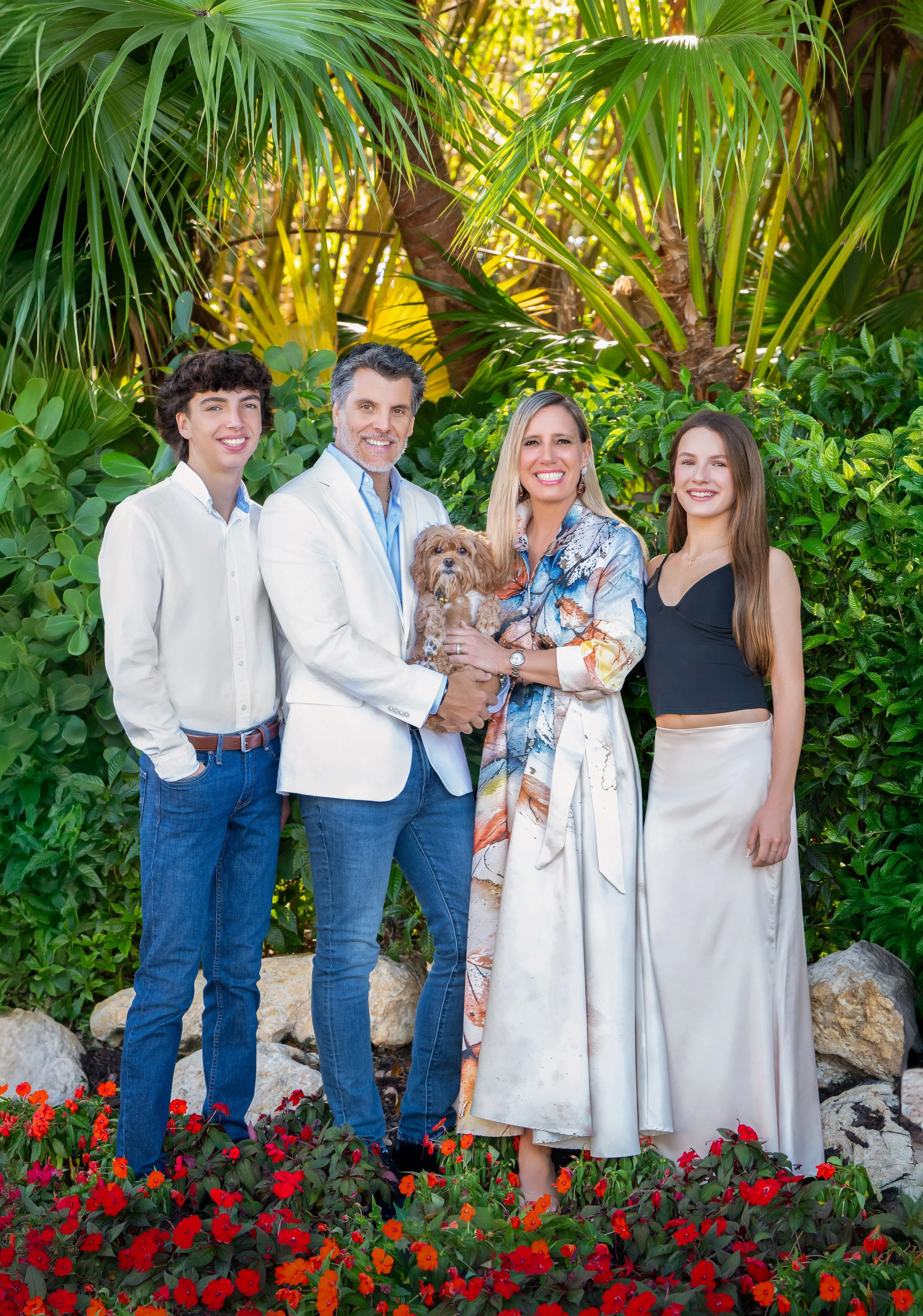 Family of four with their small dog among tropical palms and red flowers, outdoor family portrait by Vasi Studio in Palm Beach Gardens
