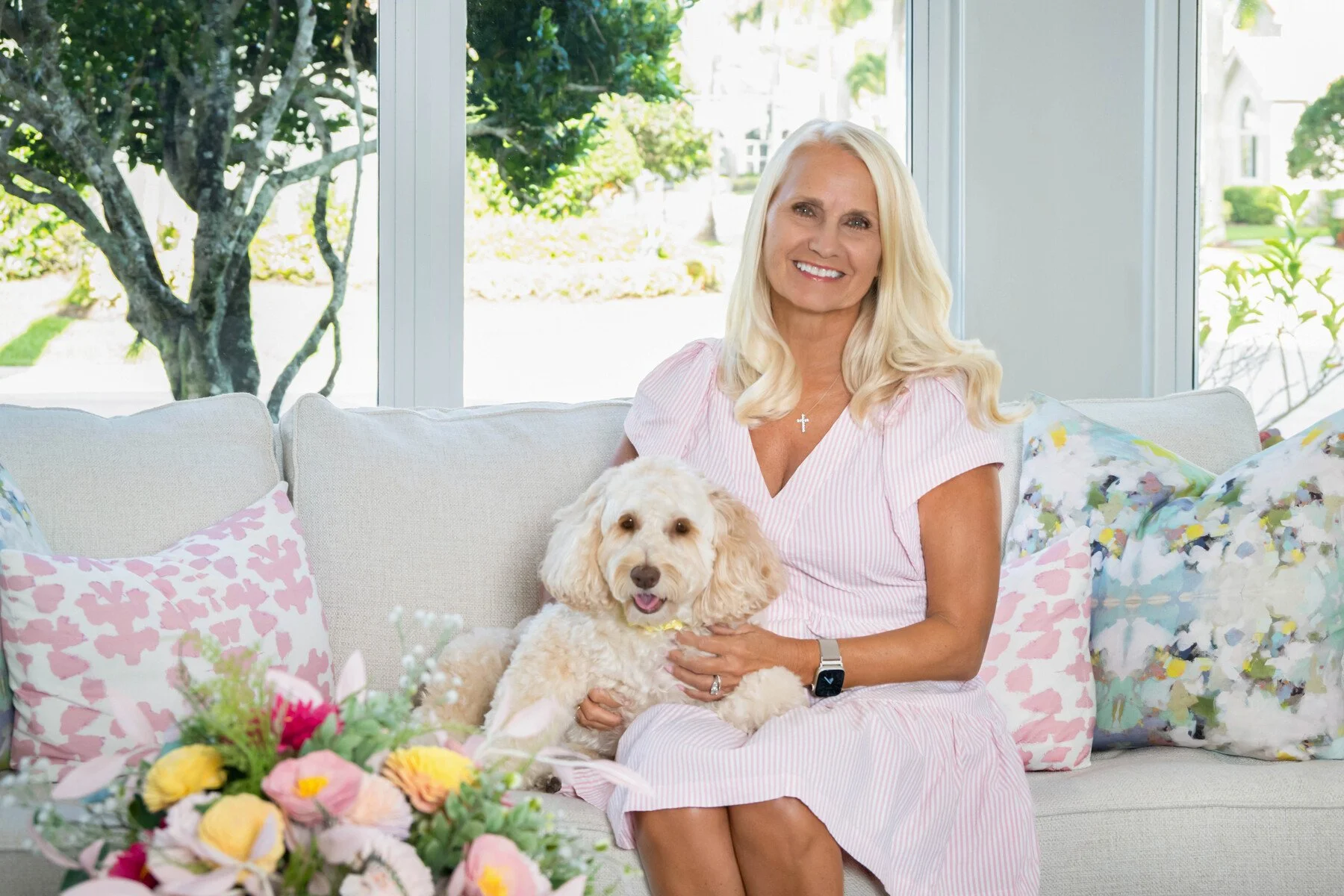 Woman with her goldendoodle on a white sofa with flowers in a bright home, in-home family portrait with pet