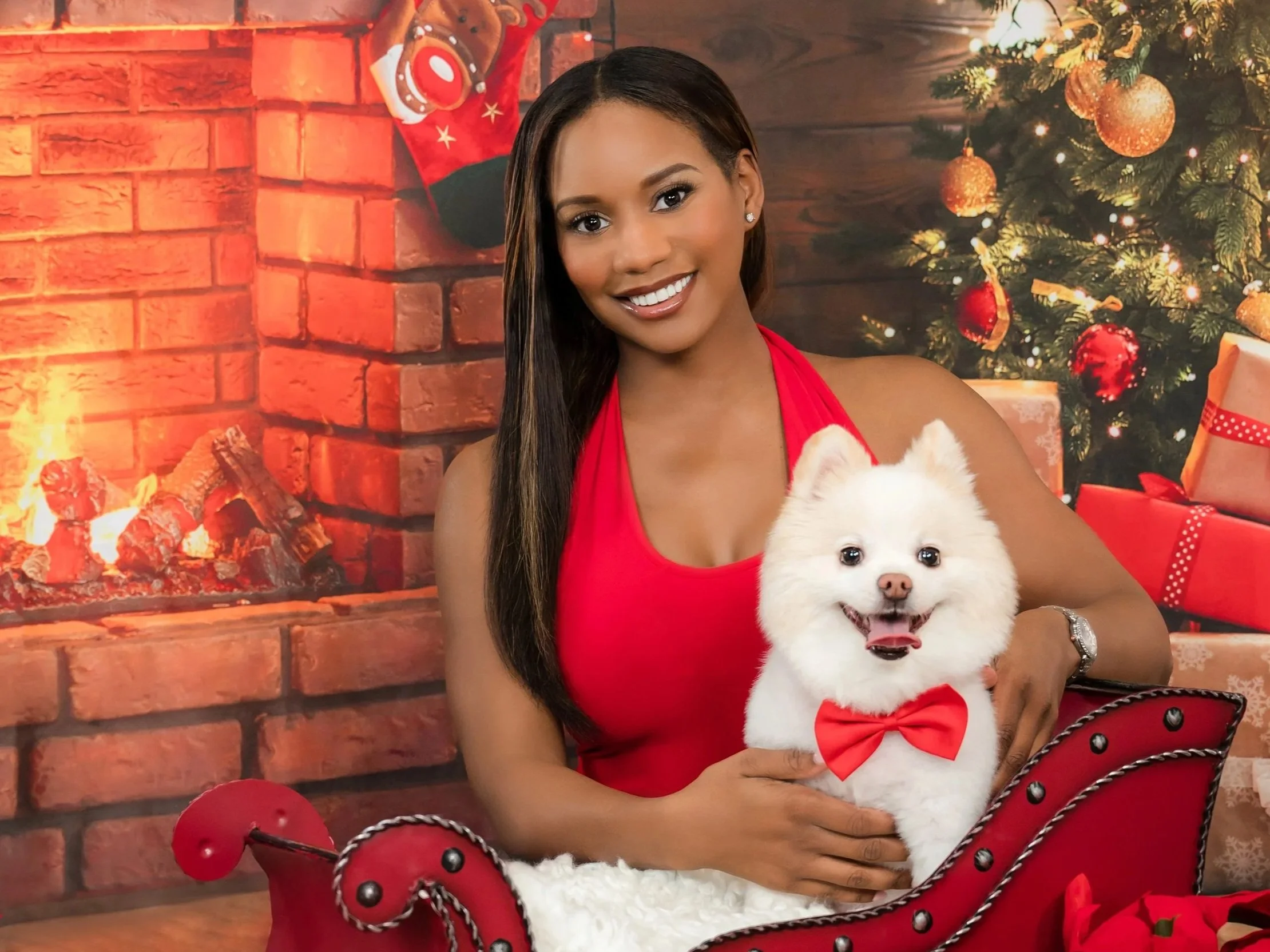 Woman in red holding a white Pomeranian with a red bow tie by a Christmas fireplace, holiday people-and-pets portrait