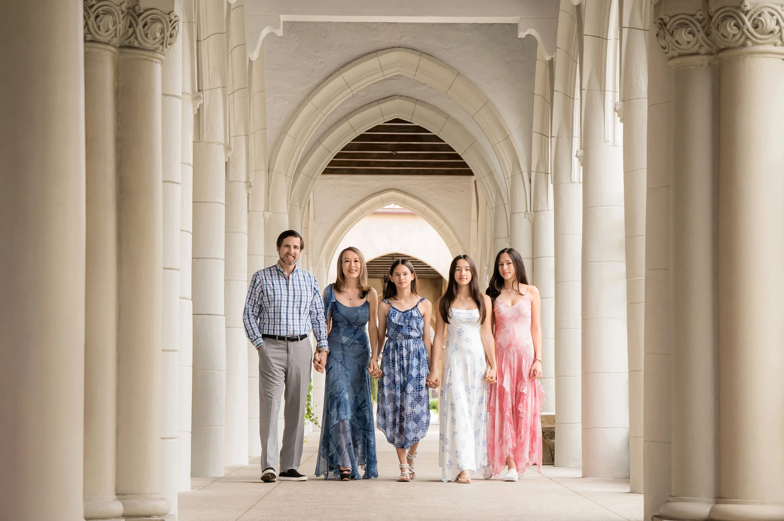 Family of five holding hands walking through an elegant white arched colonnade, outdoor family portrait by Vasi Studio, Palm Beach Gardens family photographer