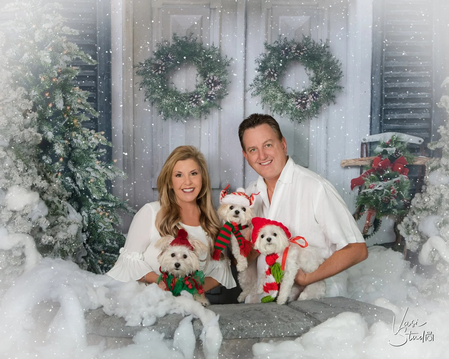 Couple with two small white dogs in Christmas sweaters in a snowy winter wreath set, holiday family and pet portrait