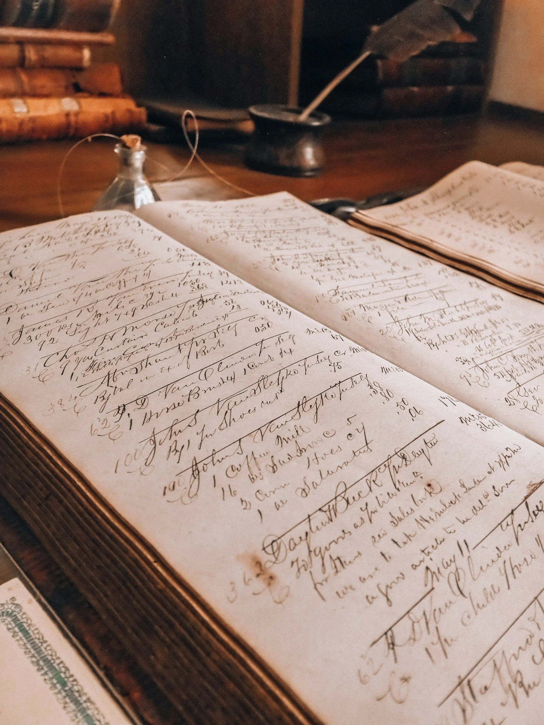Old handwritten letter or document on paper with visible cursive writing, resting on a table with a dark wooden surface, with faint background elements including a container and other objects.
