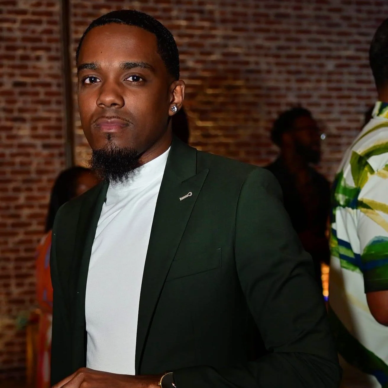 A stylish man in a dark suit with a white shirt, standing against a brick wall at an indoor event.