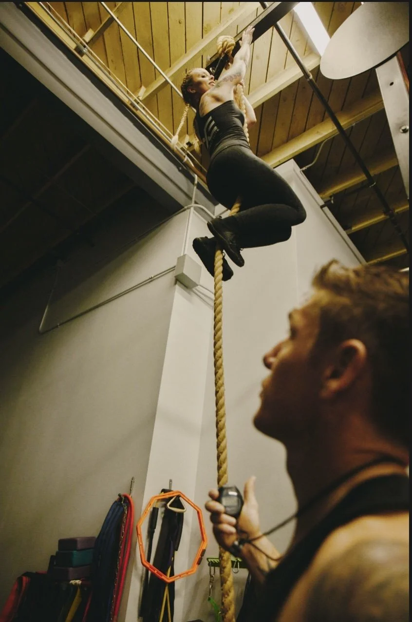 Jana climbing a rope while Daniel coaches and times her.