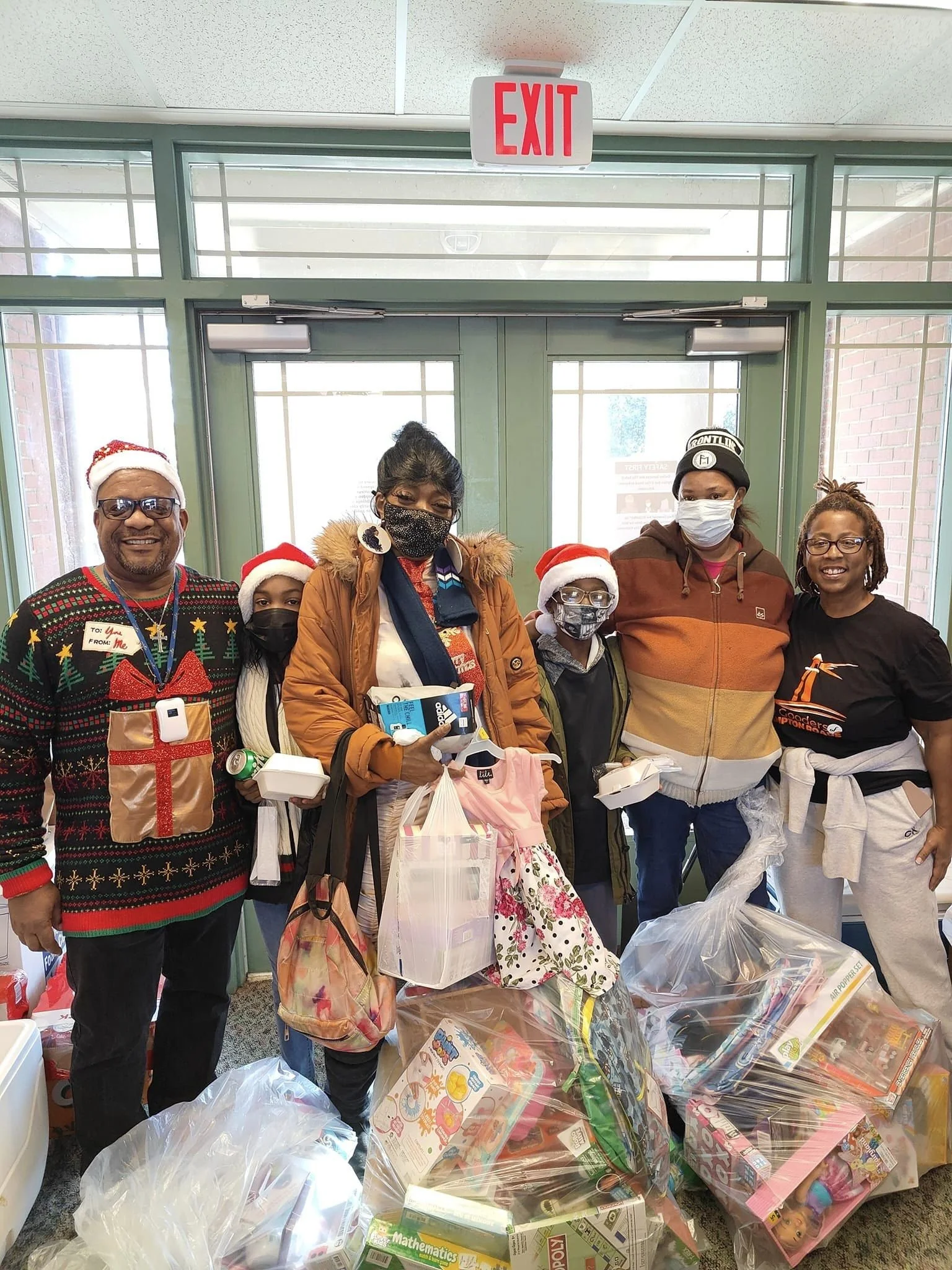 People standing inside a building beneath an exit sign, wearing Christmas hats and masks, surrounded by large bags of toys and gifts for charity.