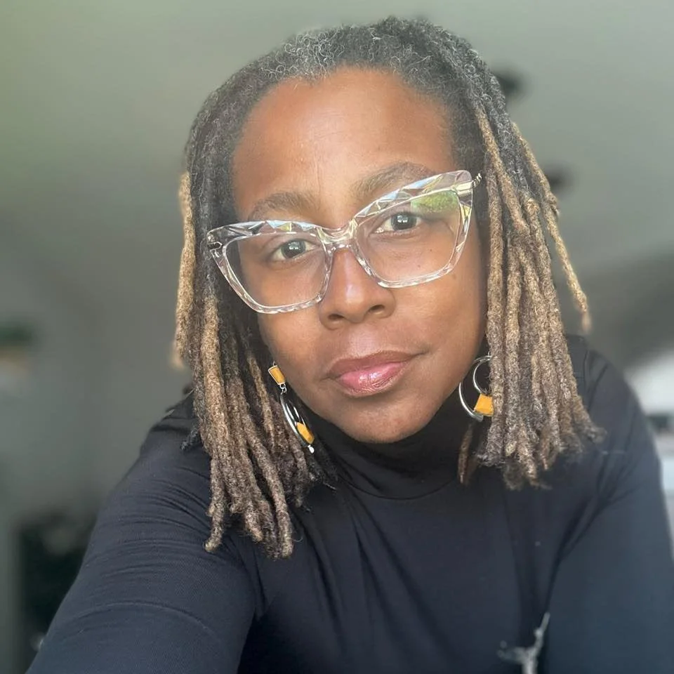 A woman with short, grayish dreadlocks wearing large, clear glasses and a black top in an indoor setting.