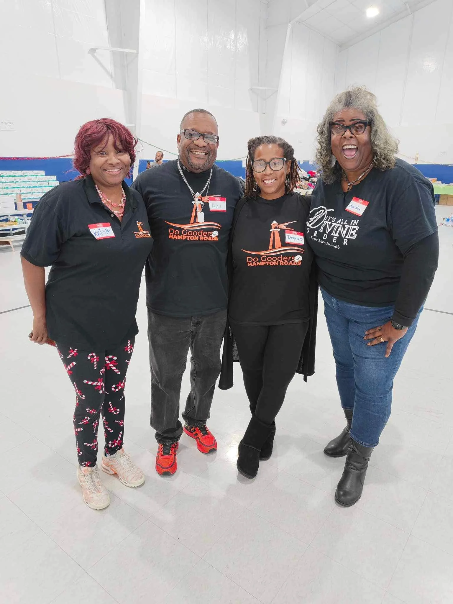 Four women standing together in a brightly lit indoor space, smiling and wearing casual clothes with name tags, with tables in the background.