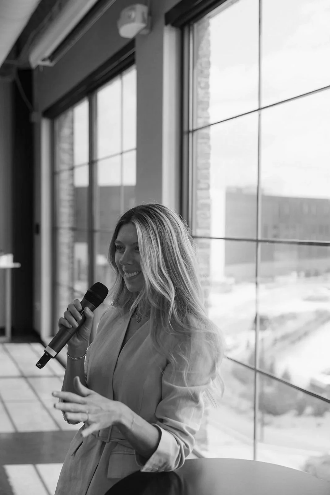 A woman with long wavy hair speaking into a microphone, standing near large windows in a modern building.